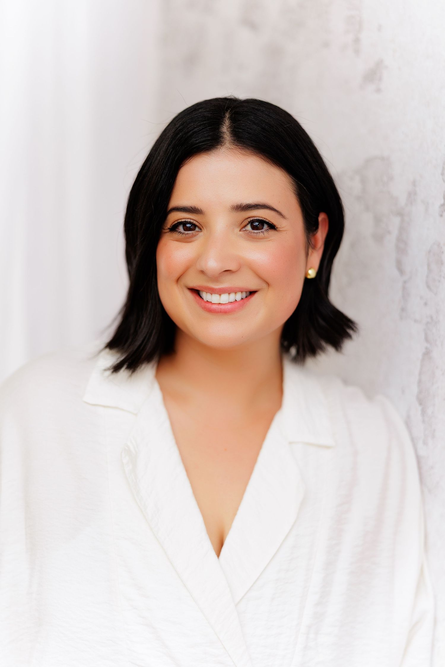Smiling woman with short dark hair wearing a white blazer poses against a light textured background.