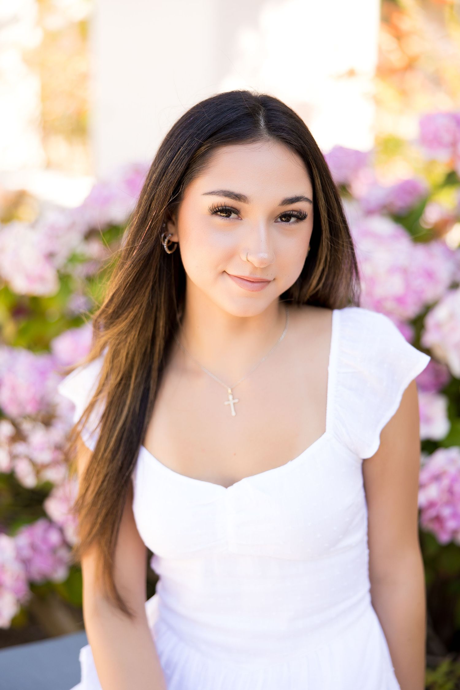 Young woman in white dress with cross necklace poses among pink hydrangeas outdoors.