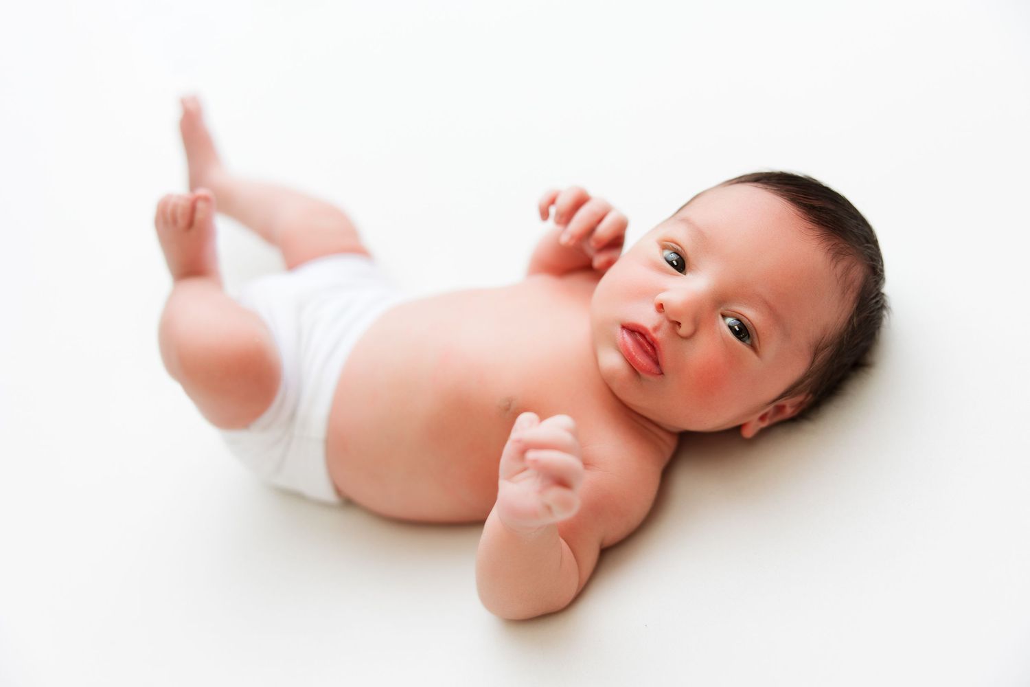 An adorable baby wearing a white diaper lies on their back on a white background, looking to the side with a content expression.