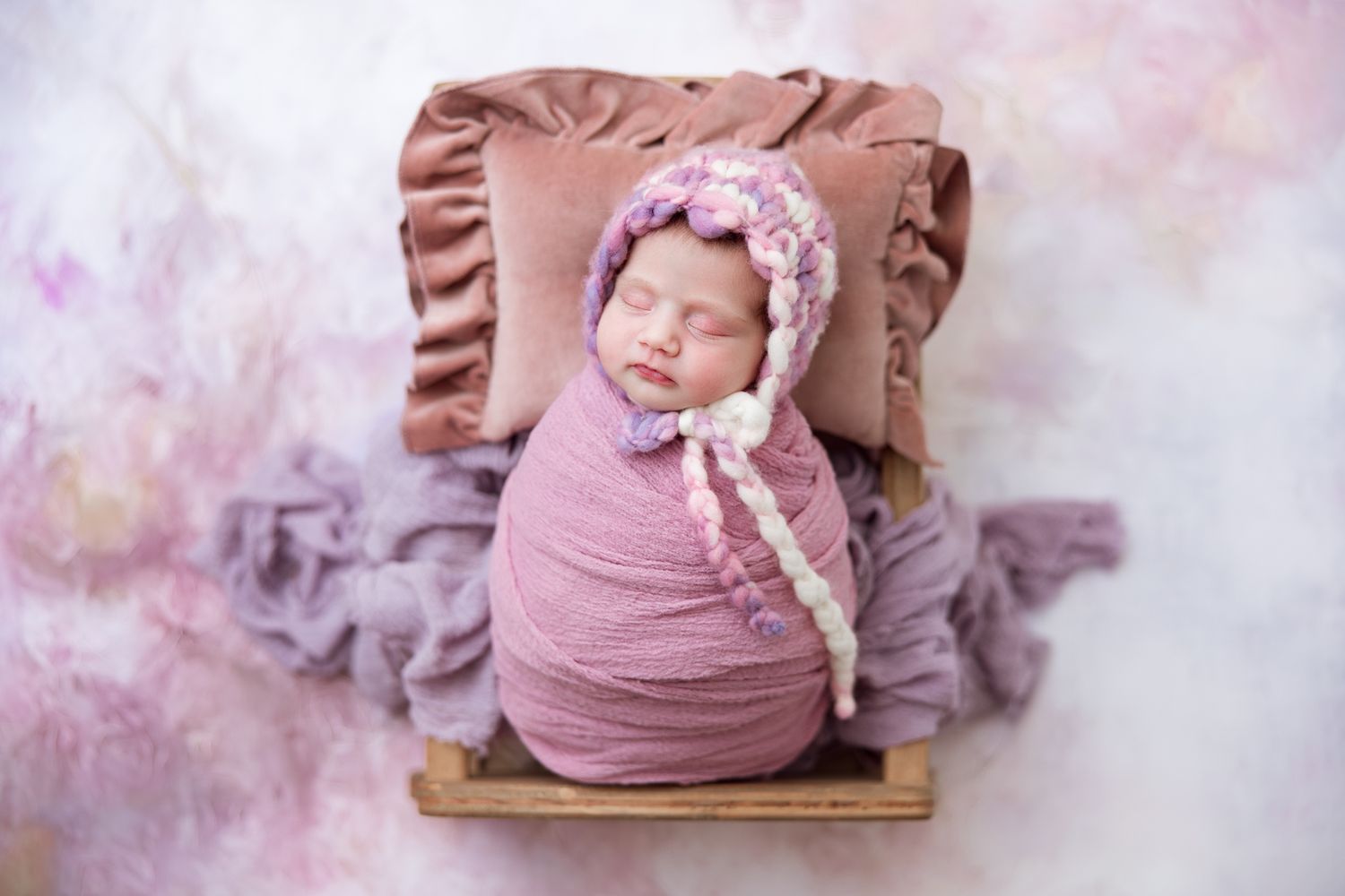 Newborn baby wearing pink swaddle blanket and headband poses during professional photoshoot with dreamy pink backdrop.