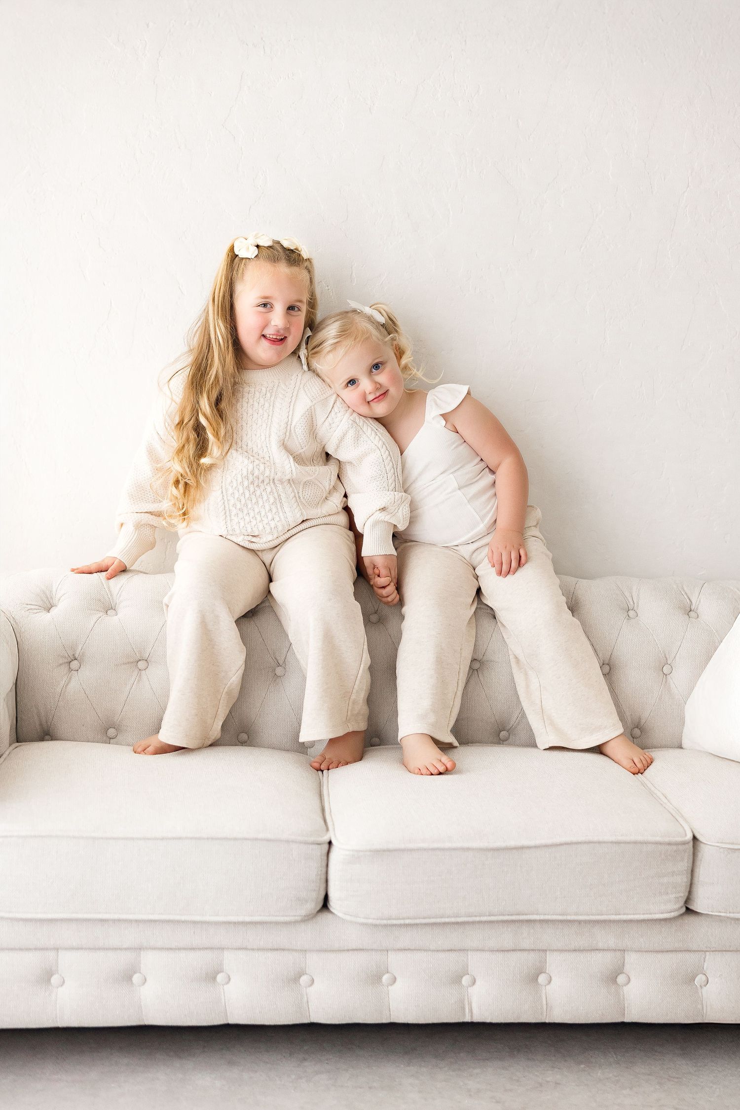 Two children in white outfits play and relax together on a plush tufted white sofa in a bright minimalist setting.