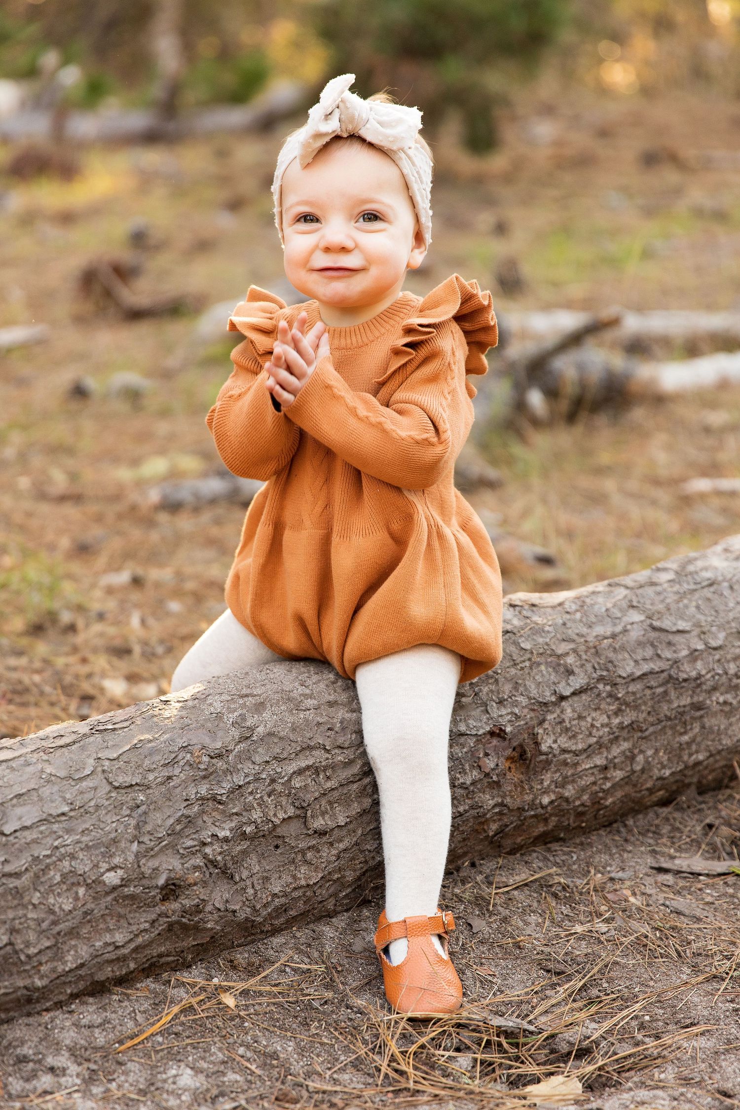 Small child sits on fallen log wearing brown dress with ruffles, white tights, and matching headband and shoes.