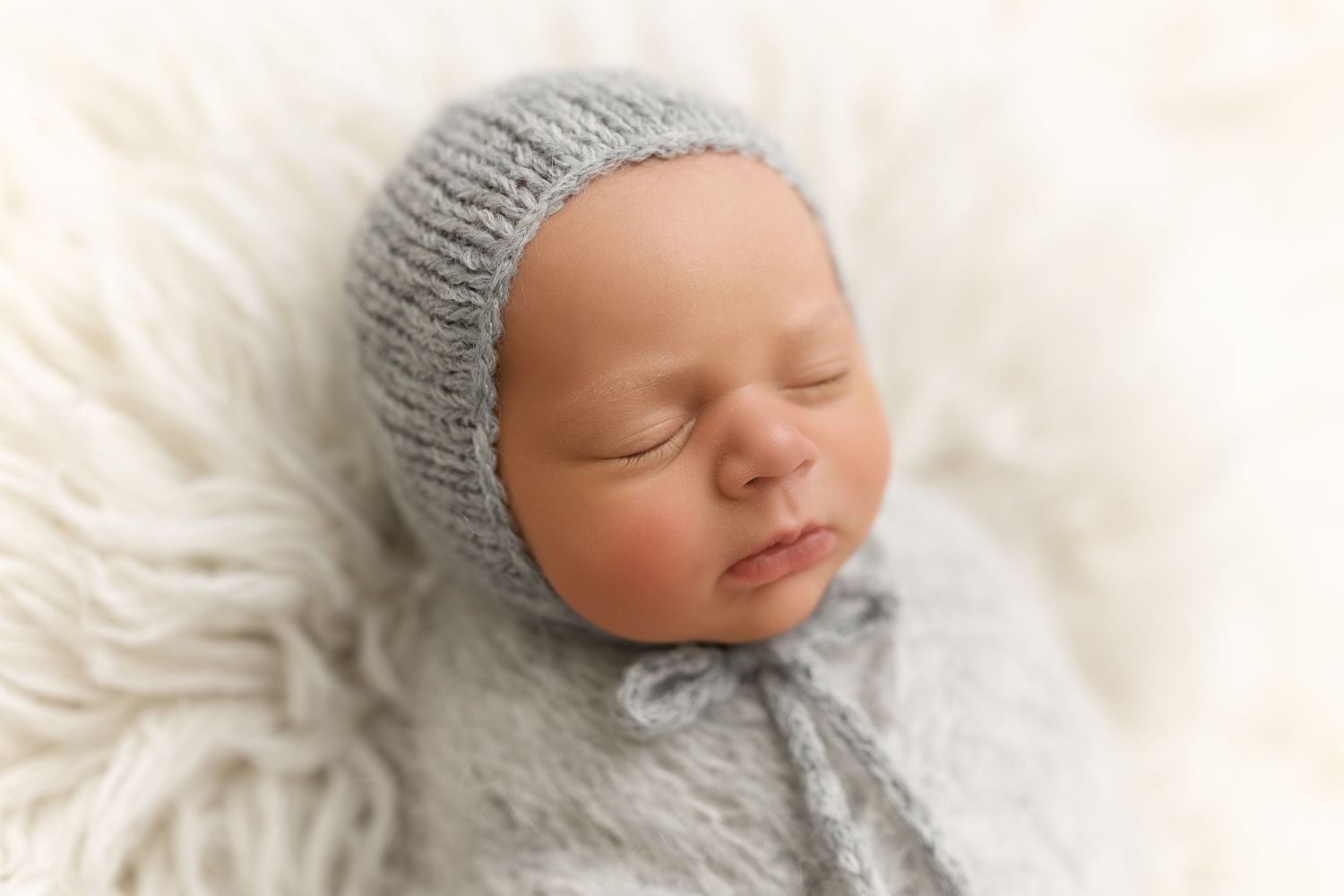 A sleeping newborn baby wearing a soft gray knit bonnet rests peacefully on white blankets.