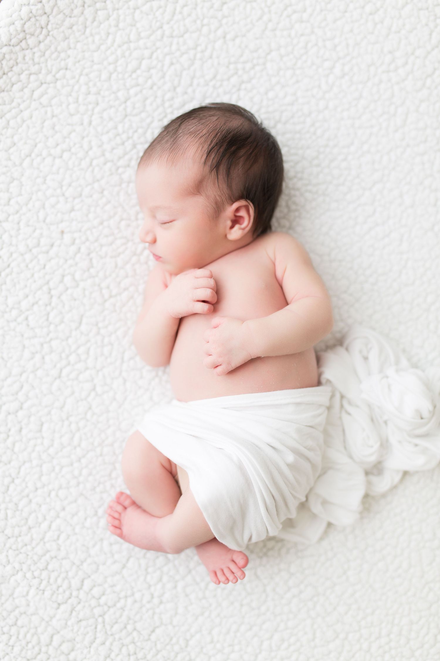 A sleeping newborn baby wrapped in white fabric lies on a textured white background.