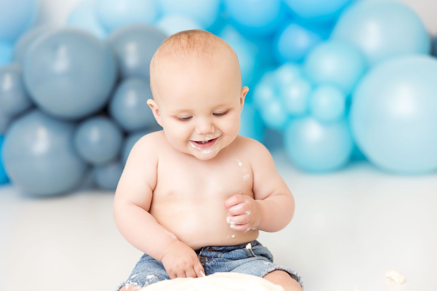 A happy baby smiles while sitting on a white floor with blue and gray balloons decorating the background.