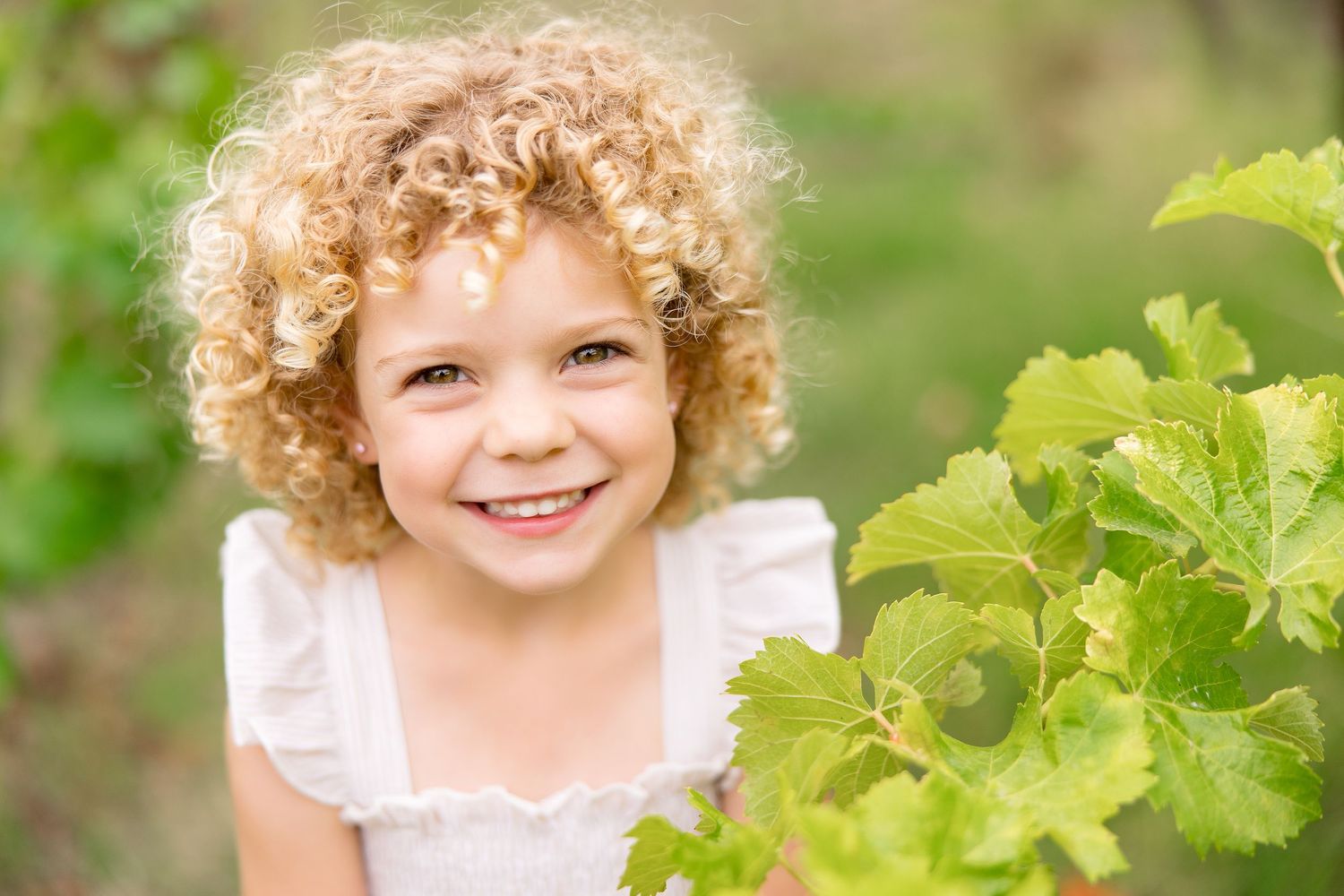 A child with curly blonde hair smiles brightly while surrounded by lush green foliage in a garden setting.