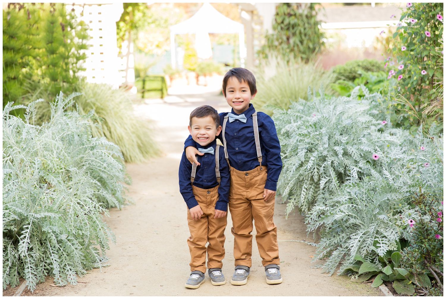 Two children wearing matching navy tops and brown pants stand together on a garden path surrounded by silvery foliage.