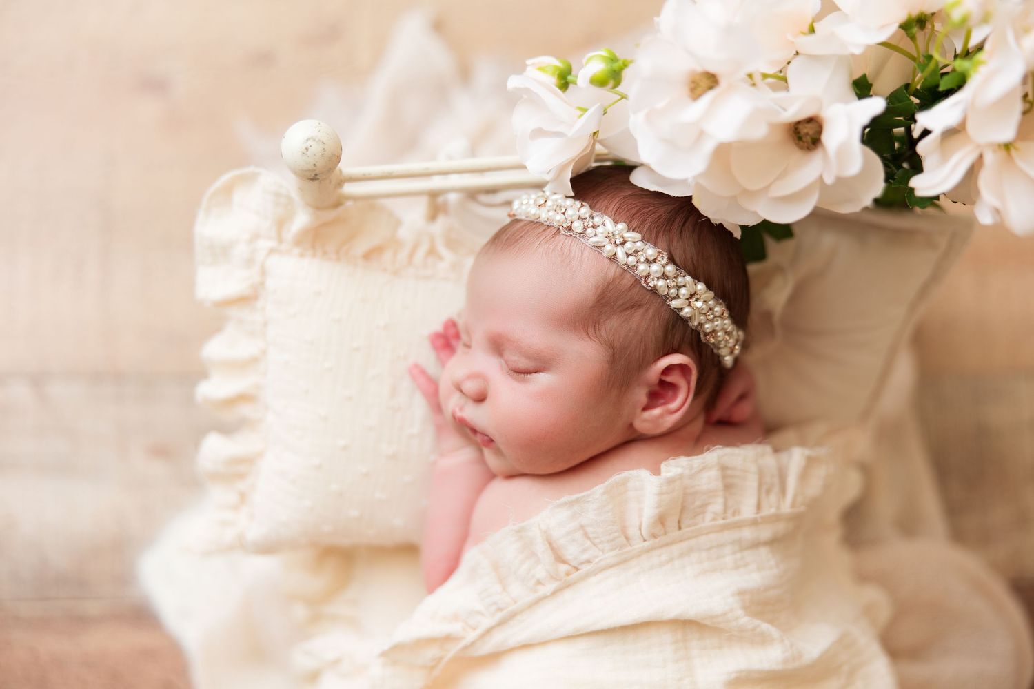 Sleeping newborn with pearl headband rests on cream pillow with floral accents.