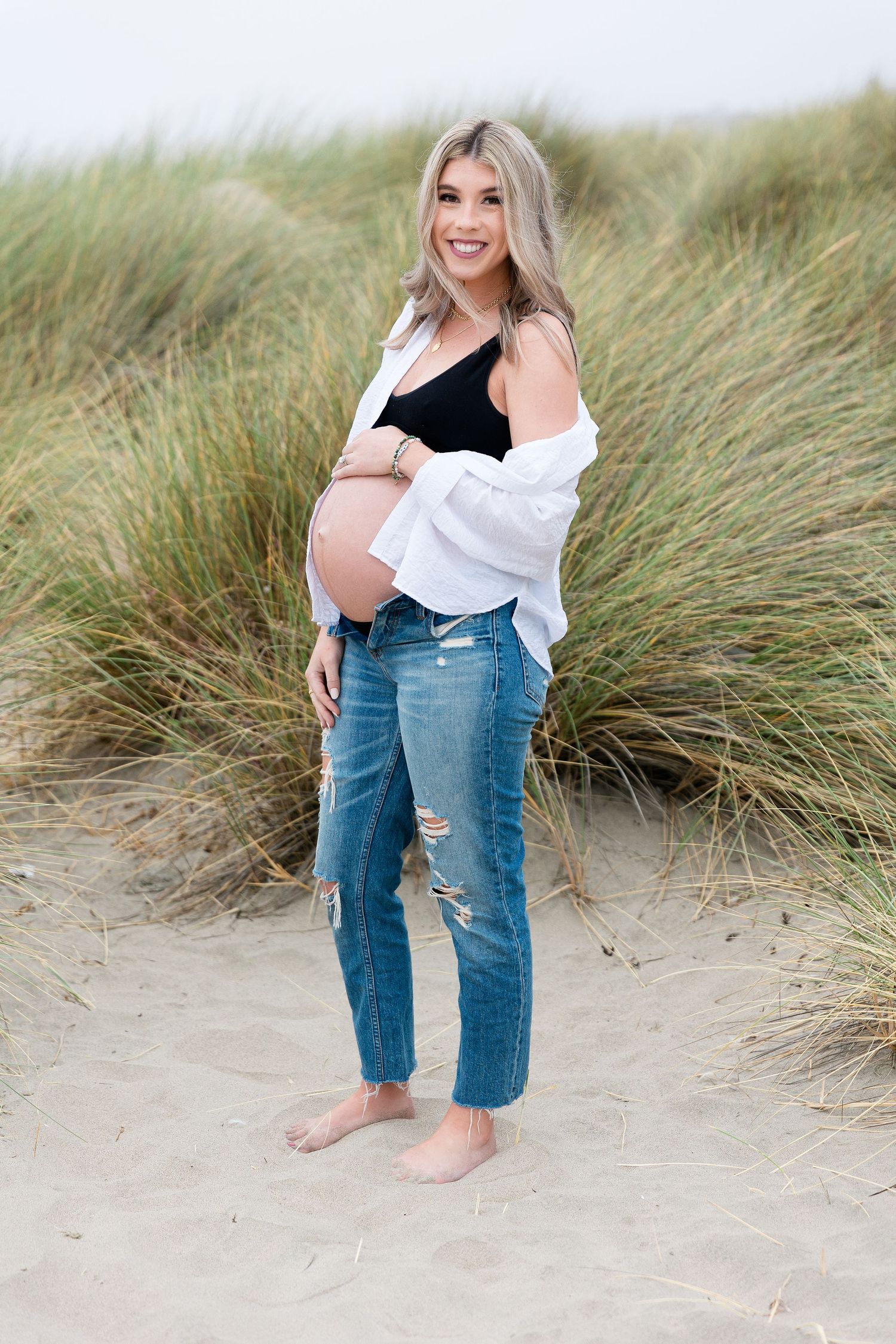 Maternity portrait of woman in distressed jeans and white cardigan among beach grass.