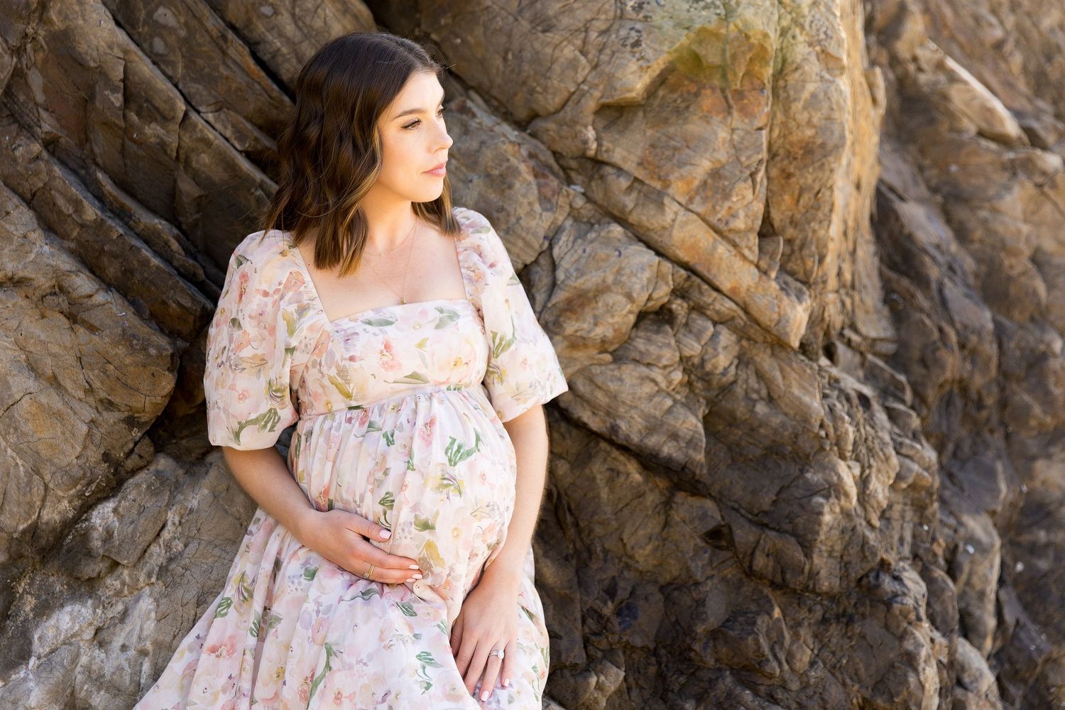 Expectant mother in floral dress poses against rocky wall background during maternity photoshoot.