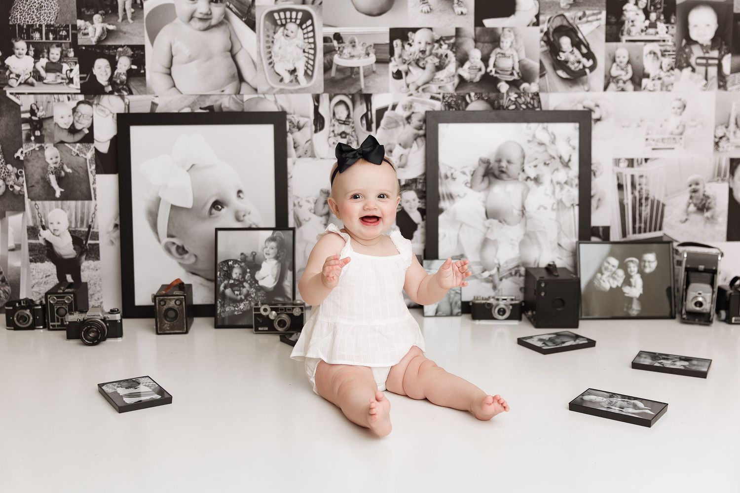 Laughing baby in white outfit sits against wall of black and white family photos.