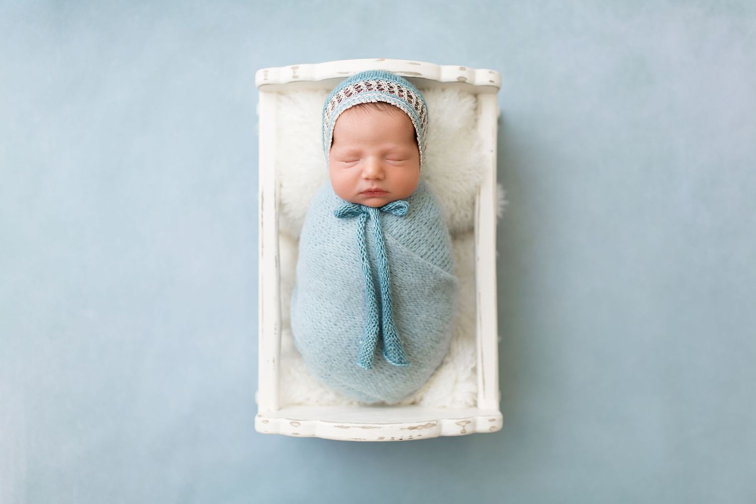 Newborn baby swaddled in blue blanket sleeping in white wooden crate.