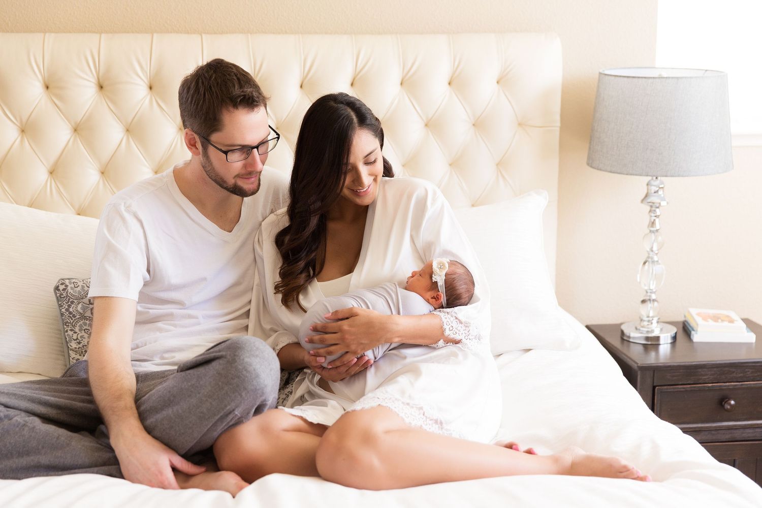 Parents cuddle with their newborn baby while sitting on a tufted headboard bed.
