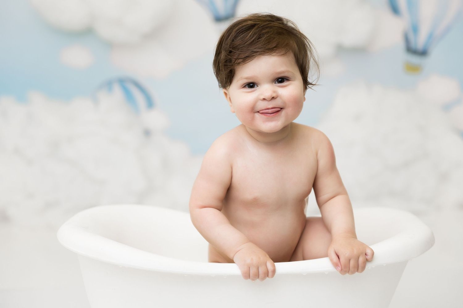 Baby sitting in white bathtub prop against dreamy cloud and hot air balloon backdrop.