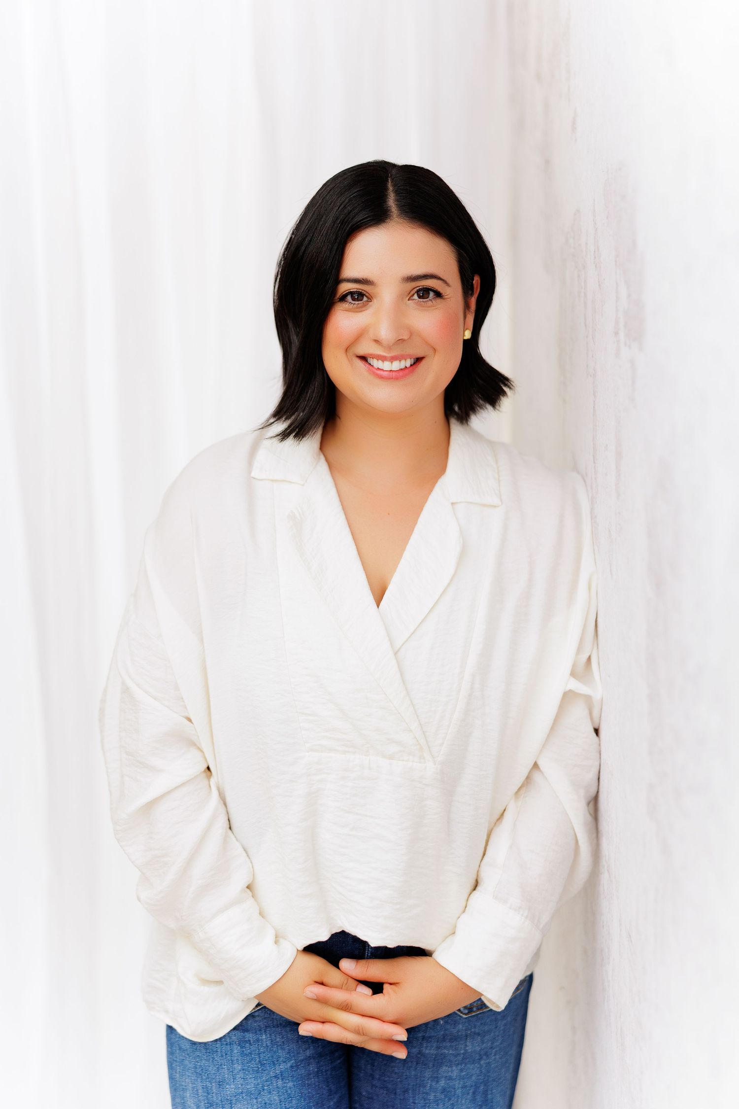 Smiling woman with dark hair wearing a white wrap blouse and jeans, standing against a white background.