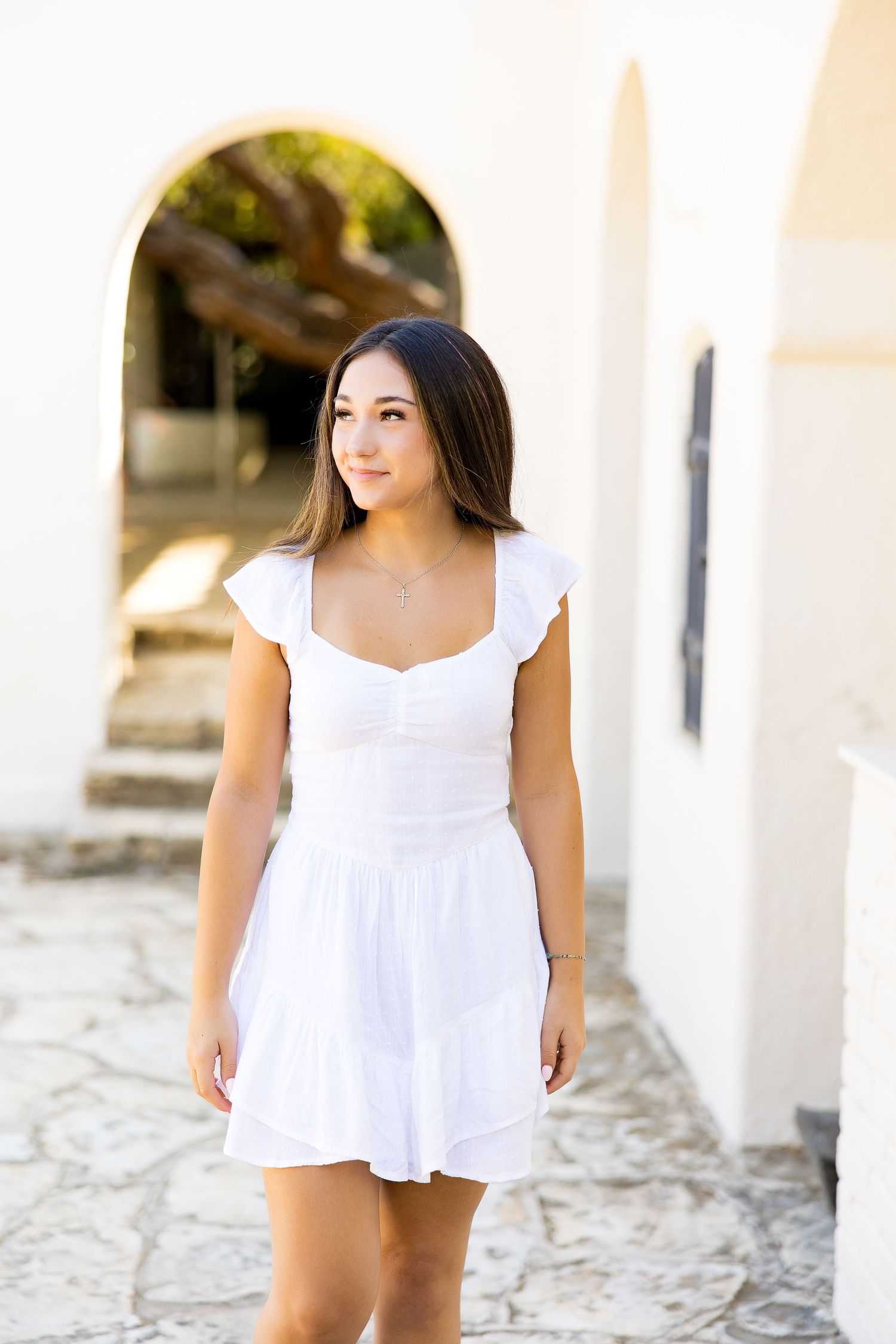 Young woman in white mini dress posing outdoors near white arched building on stone pathway.