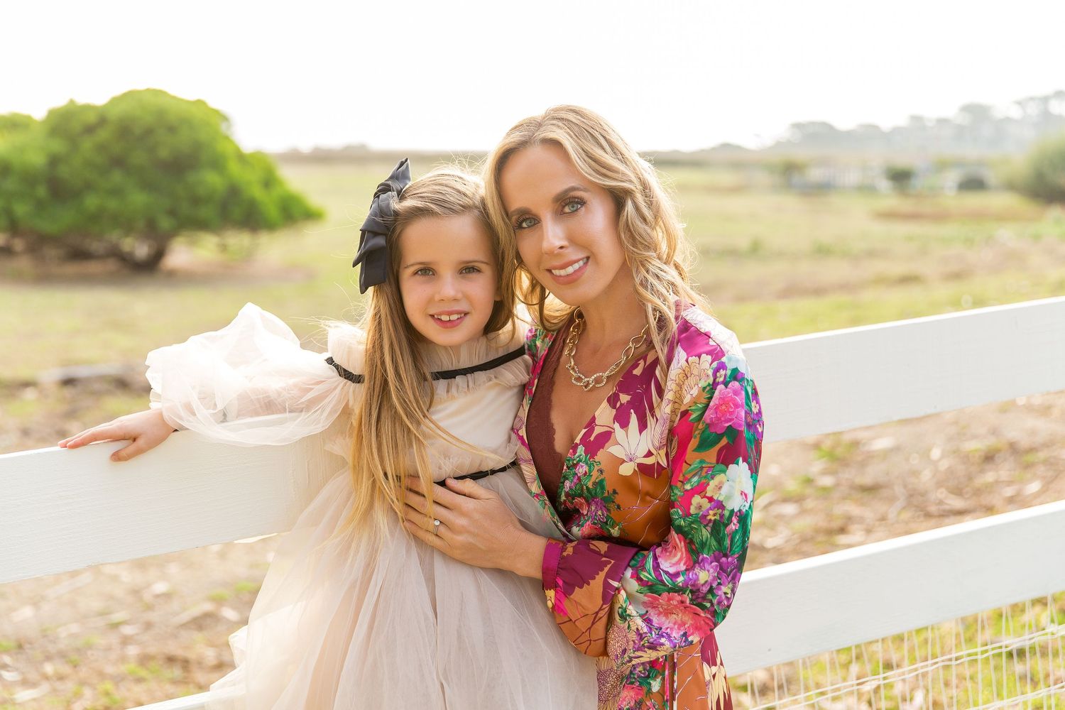 Mother and daughter in elegant dresses pose by white fence on sunny day at Carmel Vallery Ranch.