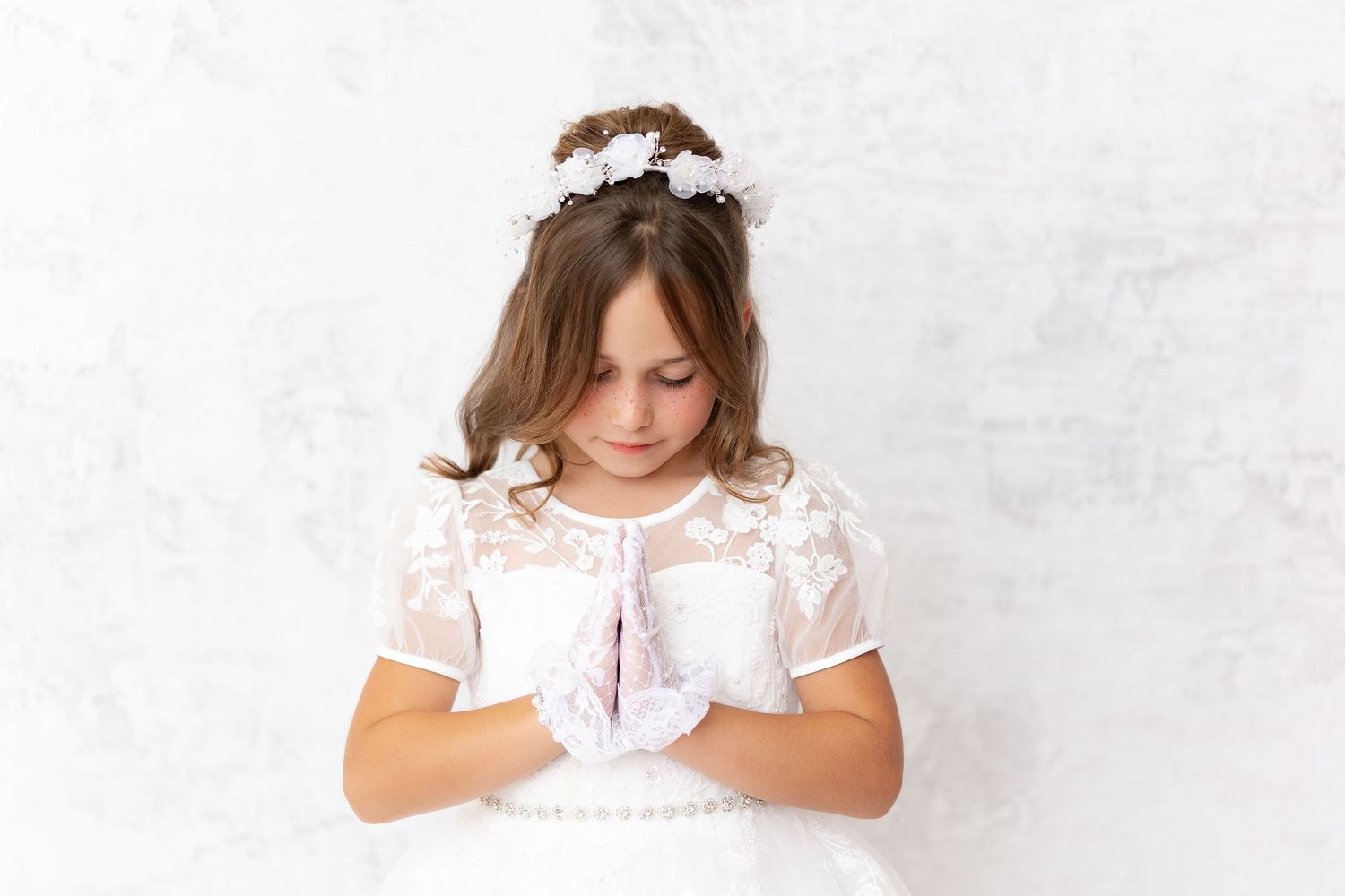 A child in a white lace dress and floral headpiece stands against a white wall looking down at their hands.