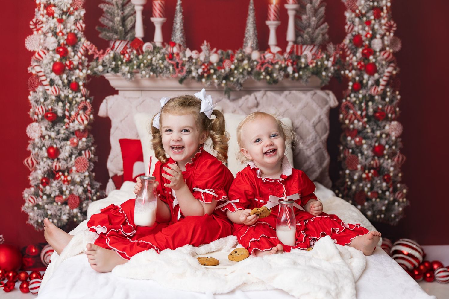 Two children in red dresses sitting on white bedding with festive Christmas decorations.
