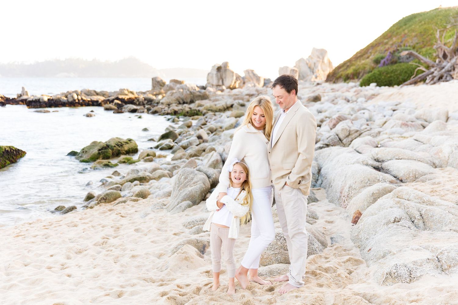 A family stands on rocky coastal shoreline at Carmel River,  at sunset with beach and ocean in the background.