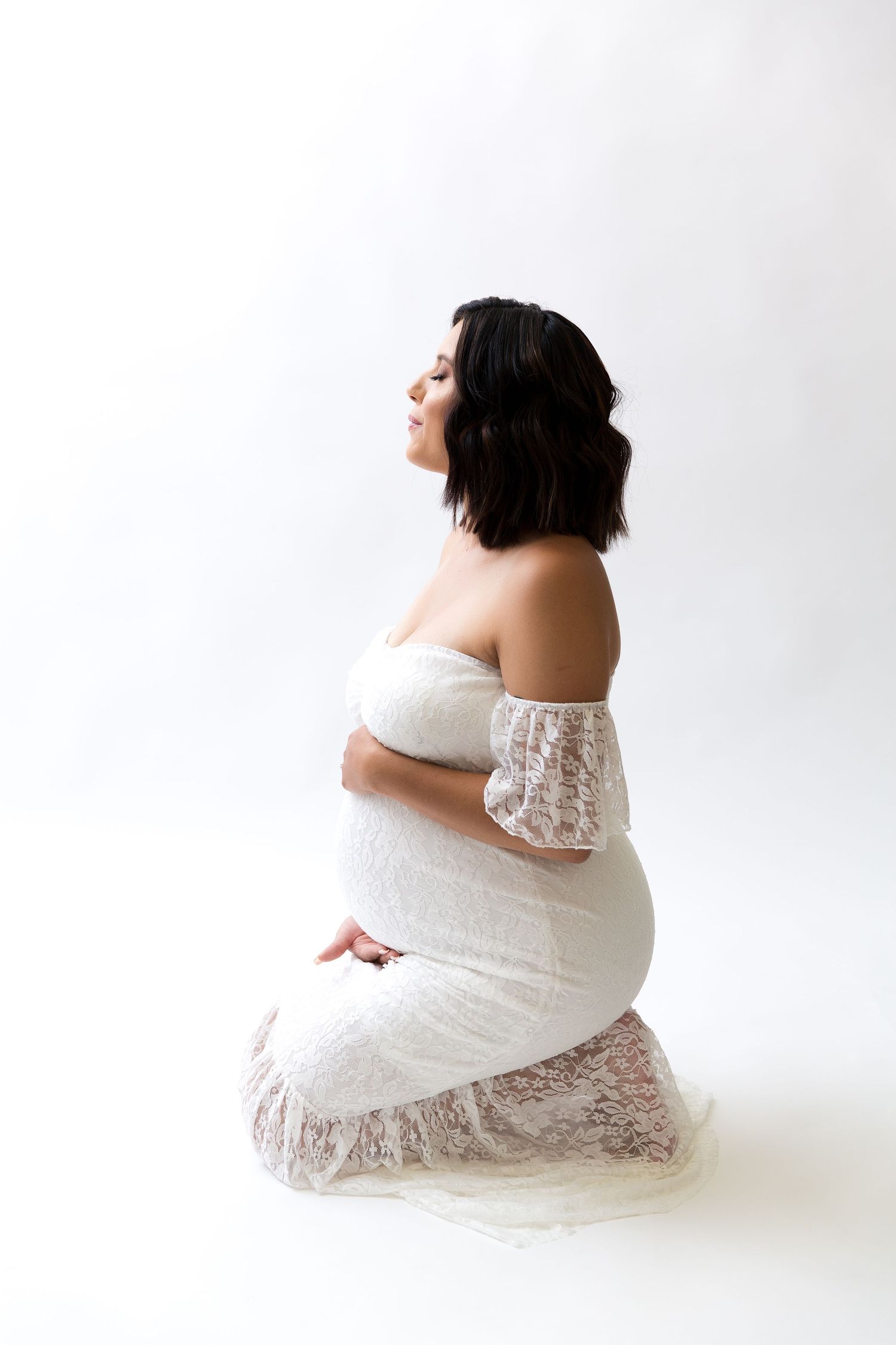 Elegant maternity portraits showing figure in white lace dress sitting against pure white backdrop.
