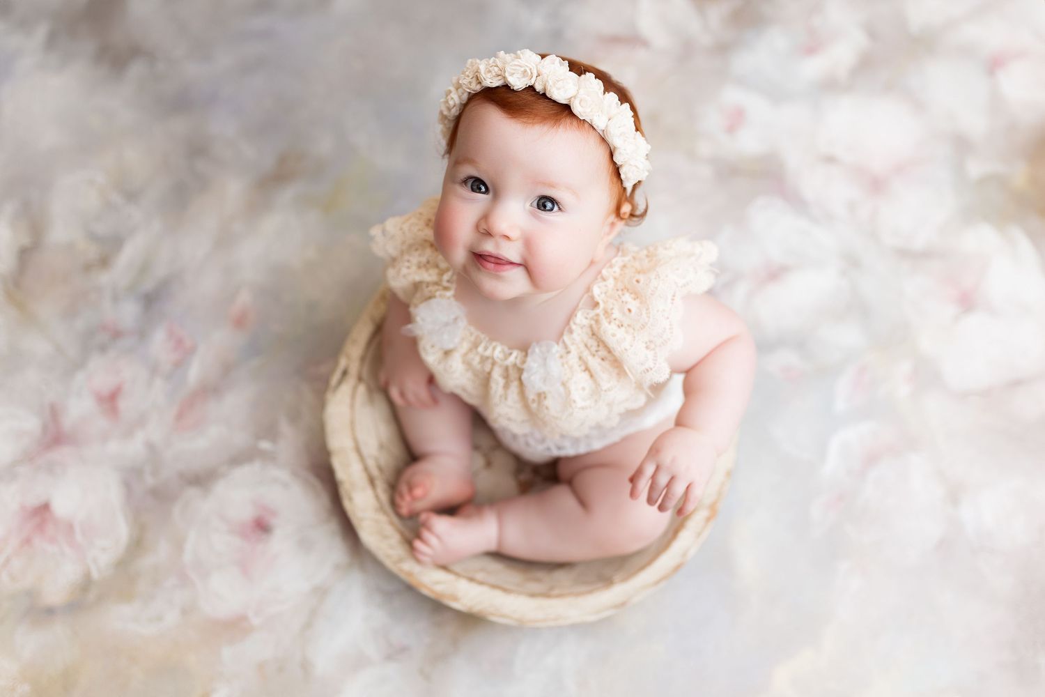 Overhead view of a baby wearing lace outfit while sitting in a wooden bowl against cream backdrop.