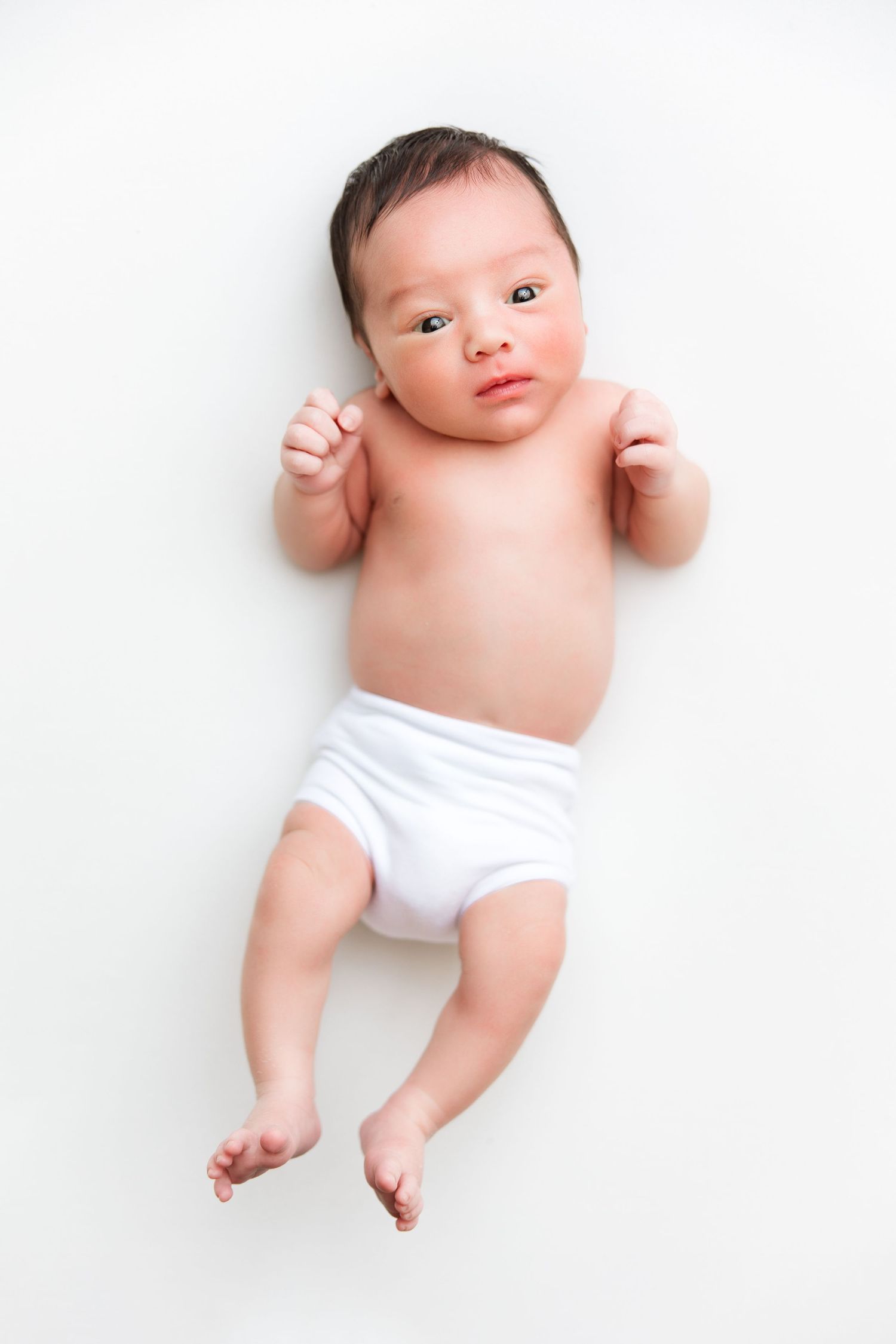 Newborn baby wearing white diaper lying on white background with tiny fists raised.