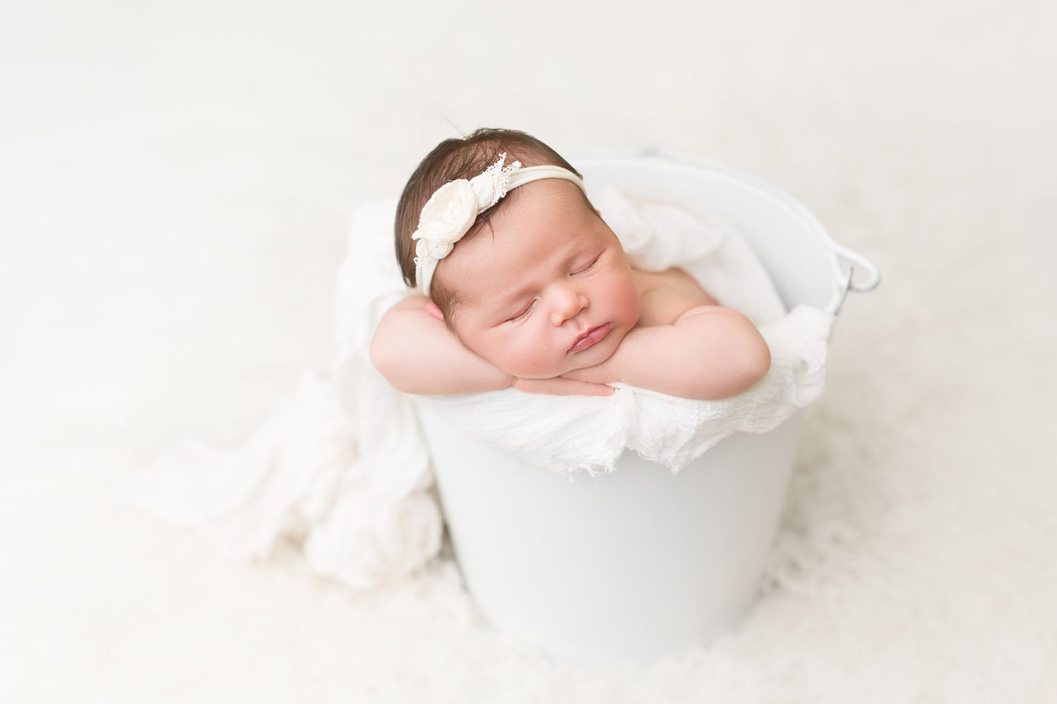 A sleeping newborn baby wrapped in white fabric lies peacefully in a white bowl.