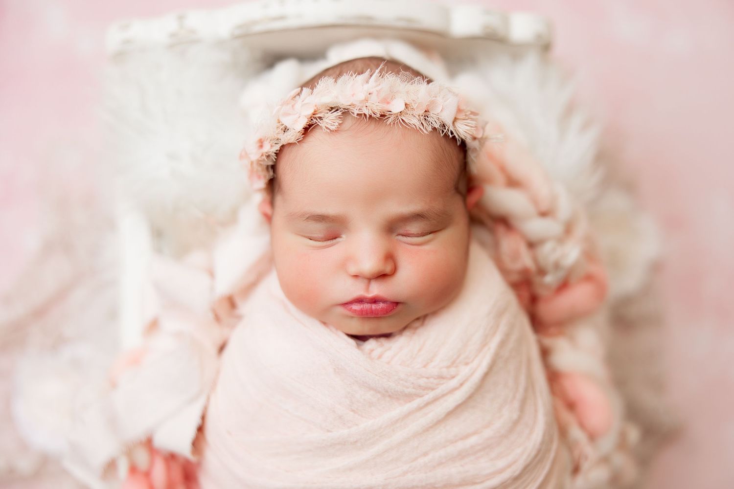 Newborn baby wearing soft pink knit hat sleeps peacefully on pink background during professional photo session.