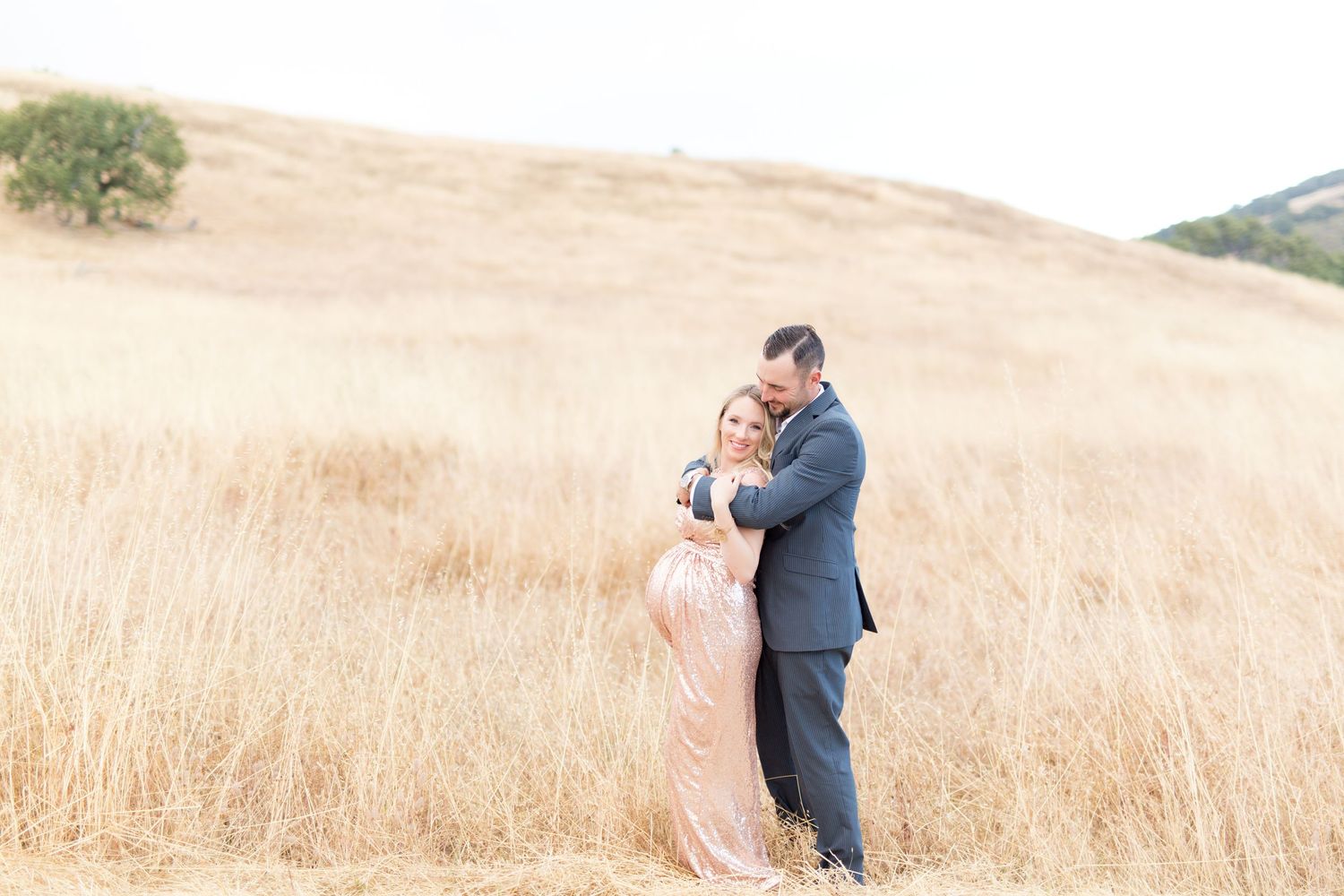 A maternity couple embraces in a golden field of tall grass during a romantic outdoor maternity photo session in Salinas.