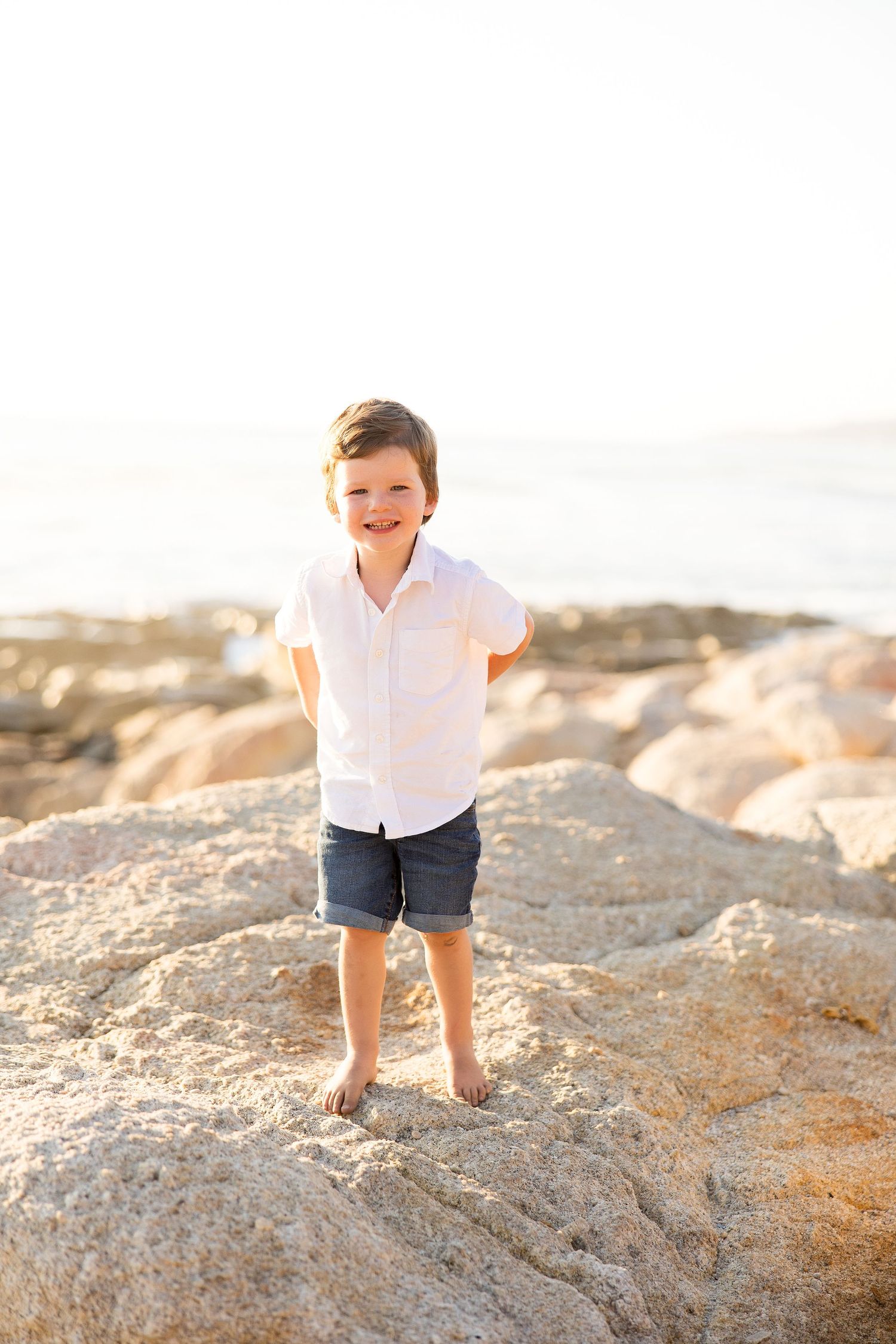 Child in white shirt and shorts playing on rocky beach during golden hour sunset.