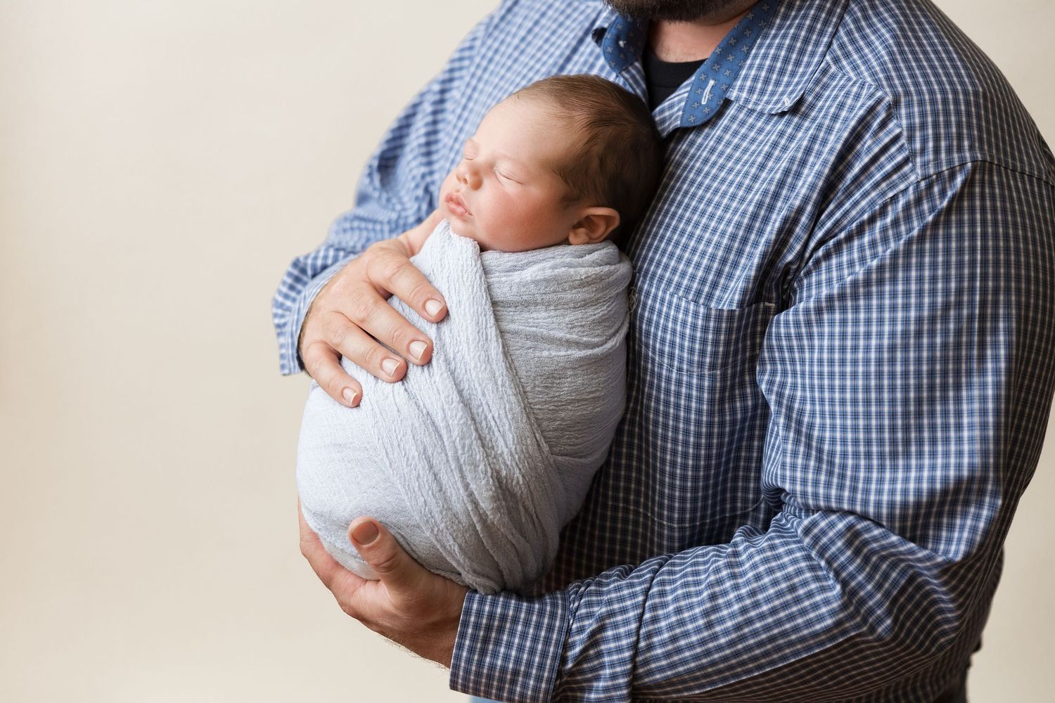Person wearing a blue checkered shirt holds sleeping baby in white onesie in nurturing embrace.