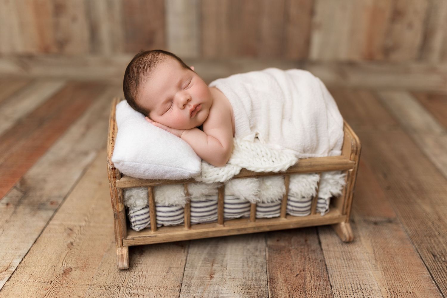 Sleeping newborn wrapped in white swaddle lying on rustic wooden surface and cushions.