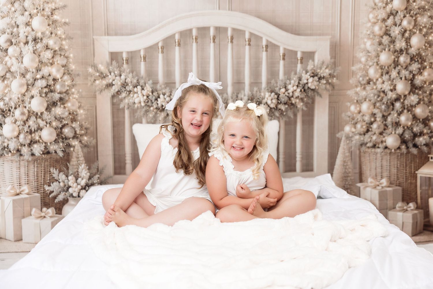 Two young girls in white dresses and bows sit smiling on a white bed surrounded by flocked Christmas trees and wrapped gifts.