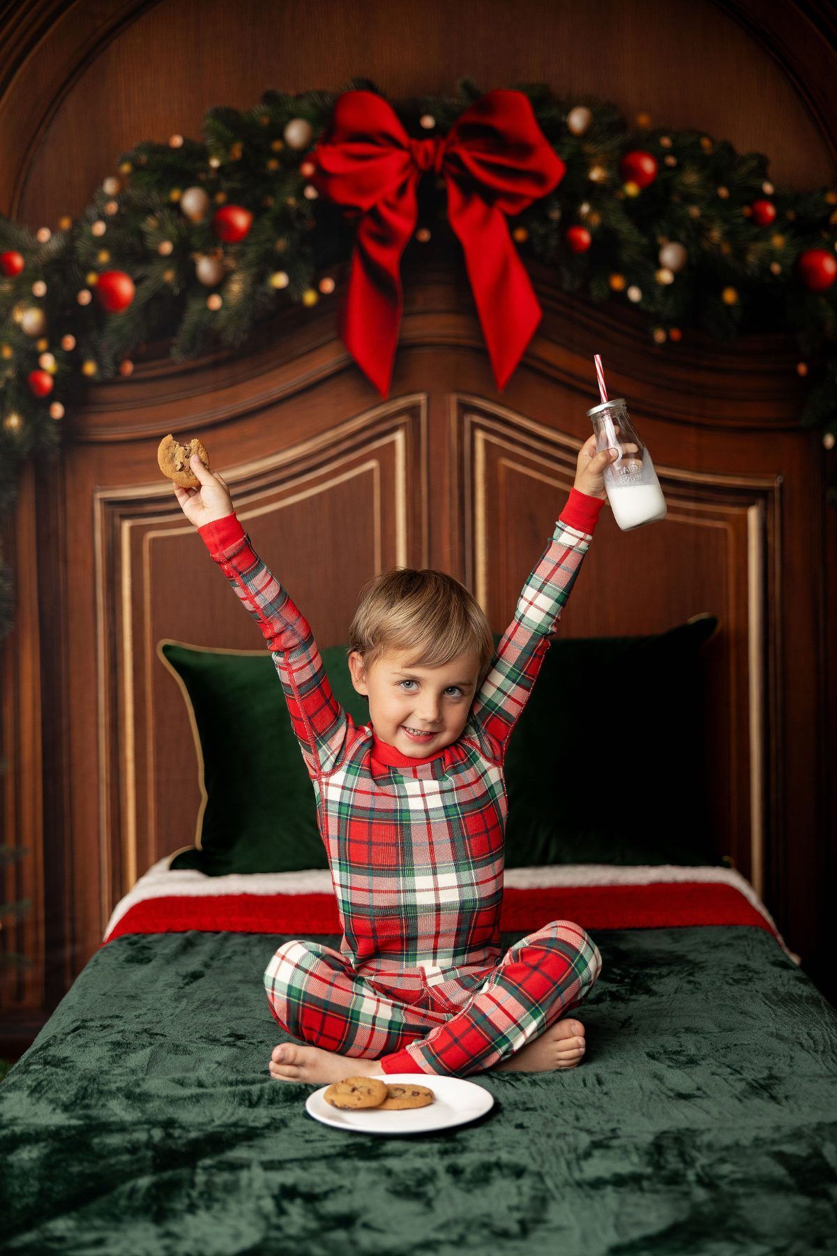 Boy in plaid Christmas pajamas cheering with milk and cookies on a cozy Christmas bed set