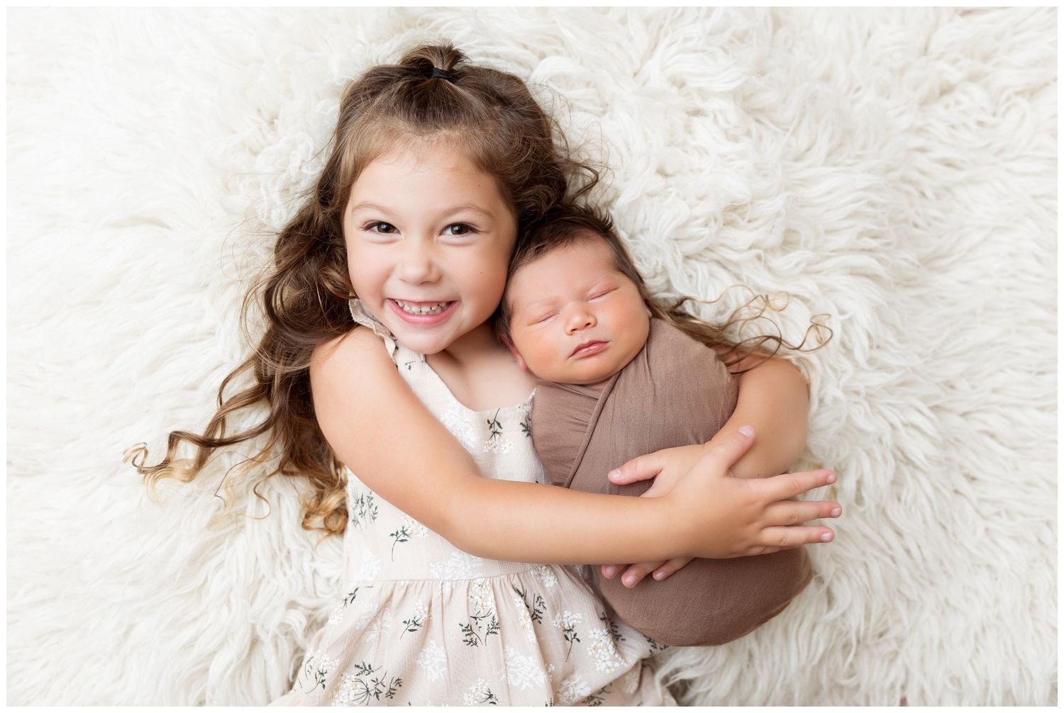 Two young children in white and brown outfits lay together on a soft white fur blanket.
