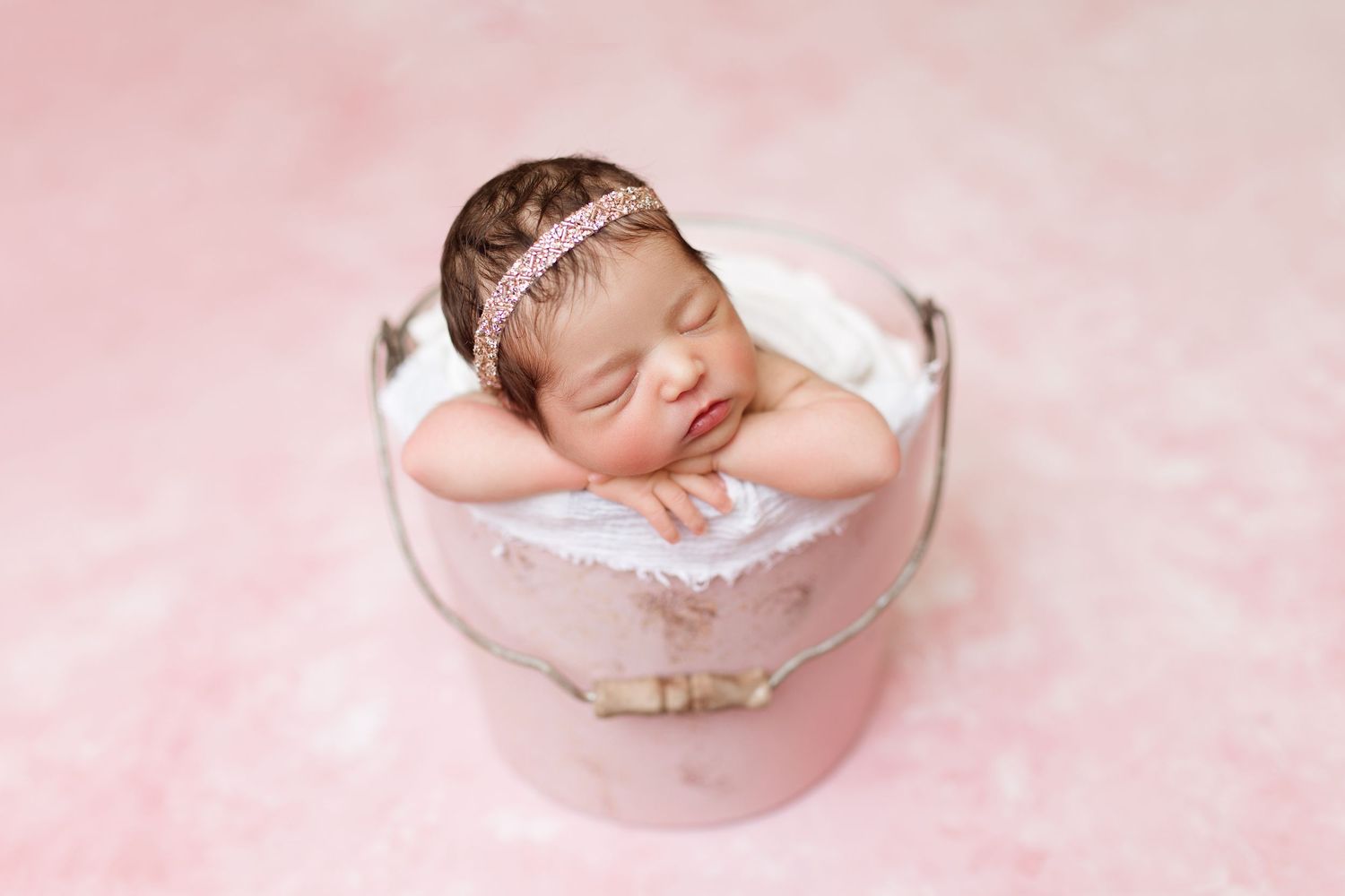 Sleeping newborn wearing delicate headband nestled in pink ceramic bowl on soft pink background.