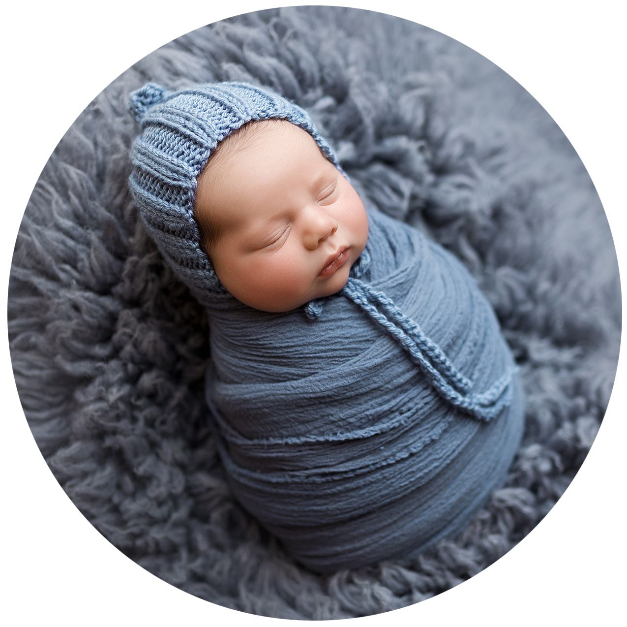 Newborn baby sleeping peacefully wrapped in soft gray swaddle blanket on plush gray fur backdrop.