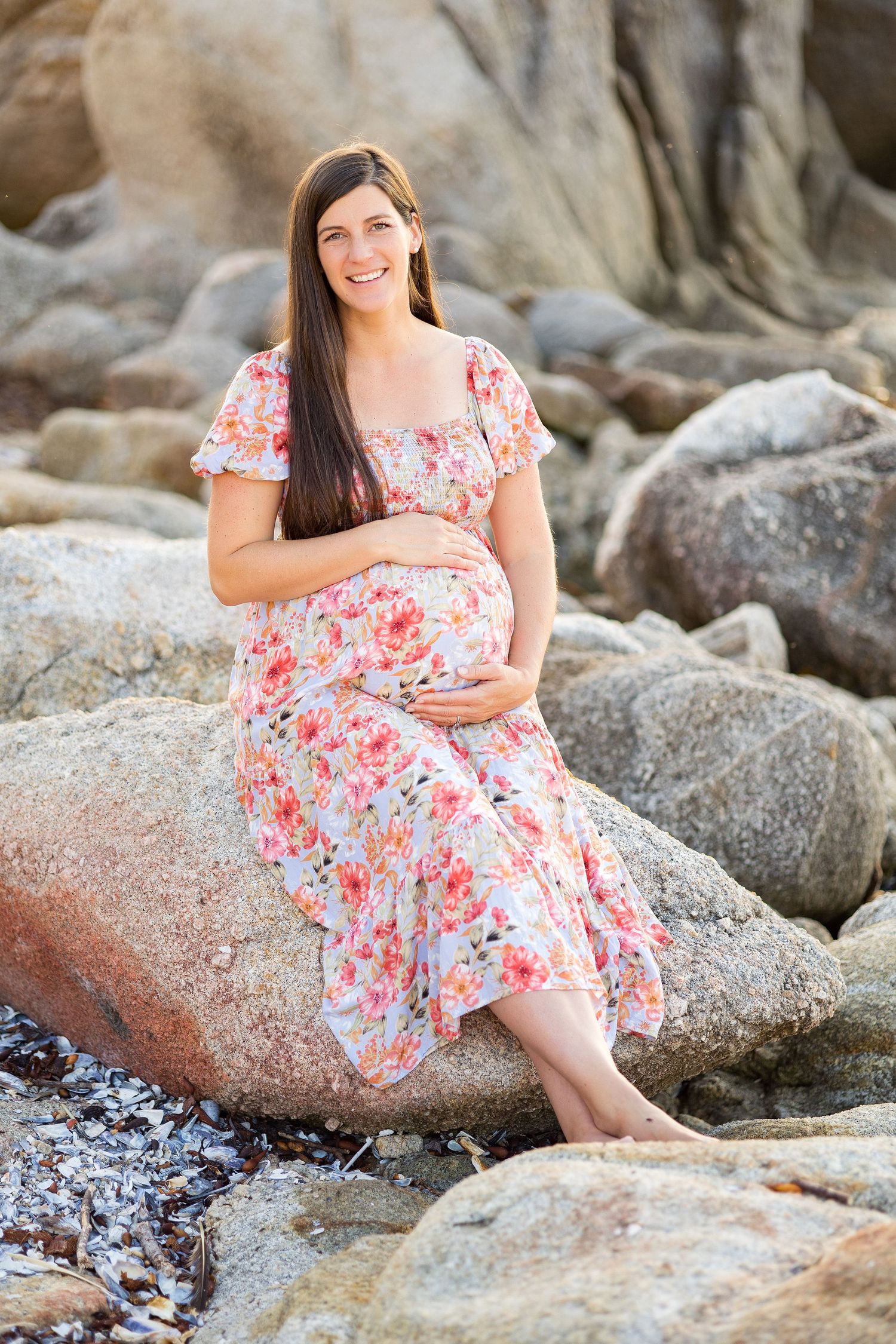 Woman in floral dress sitting on coastal rocks during maternity photoshoot.