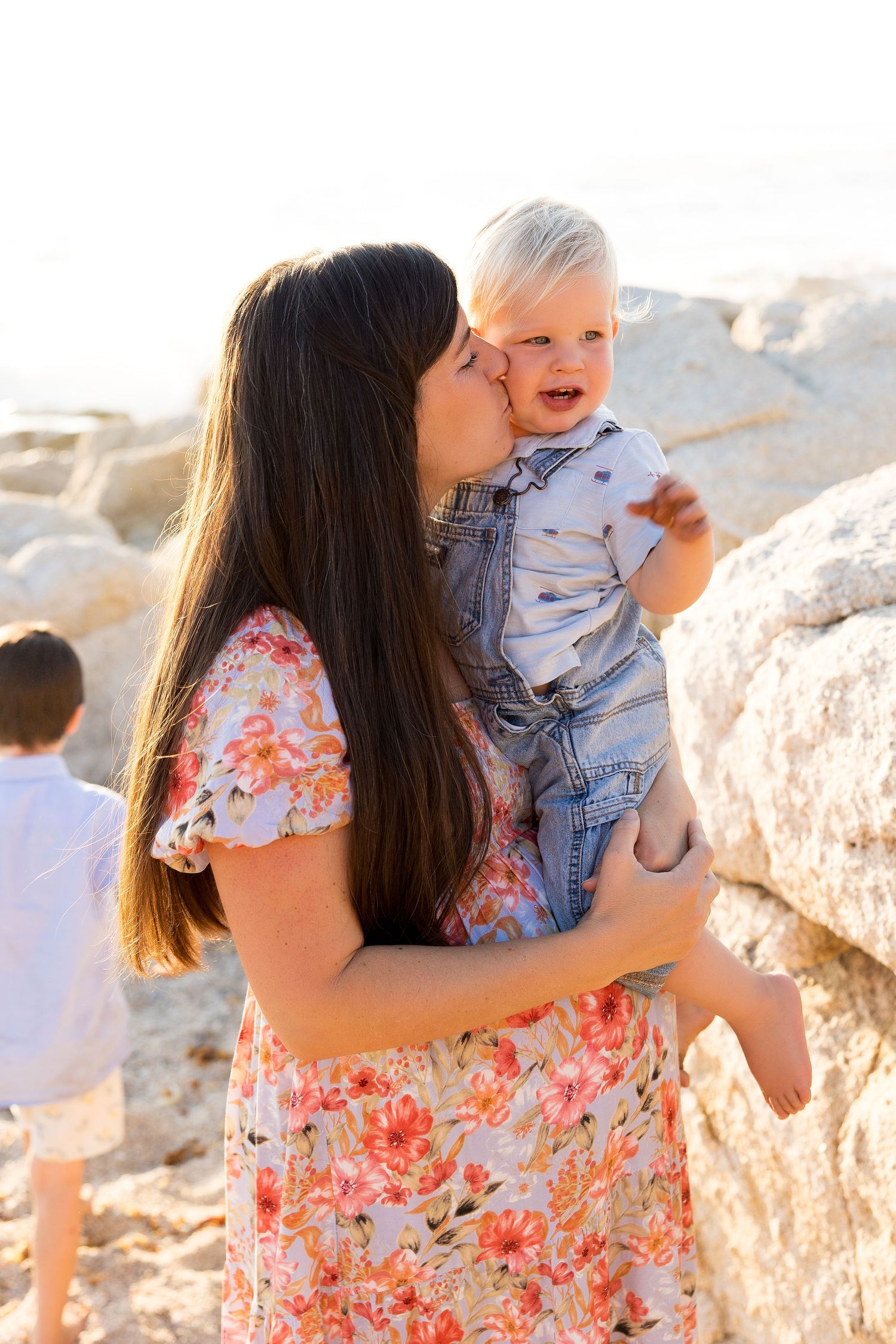 A floral dress-clad woman holds and kisses a baby among rocky coastal scenery.
