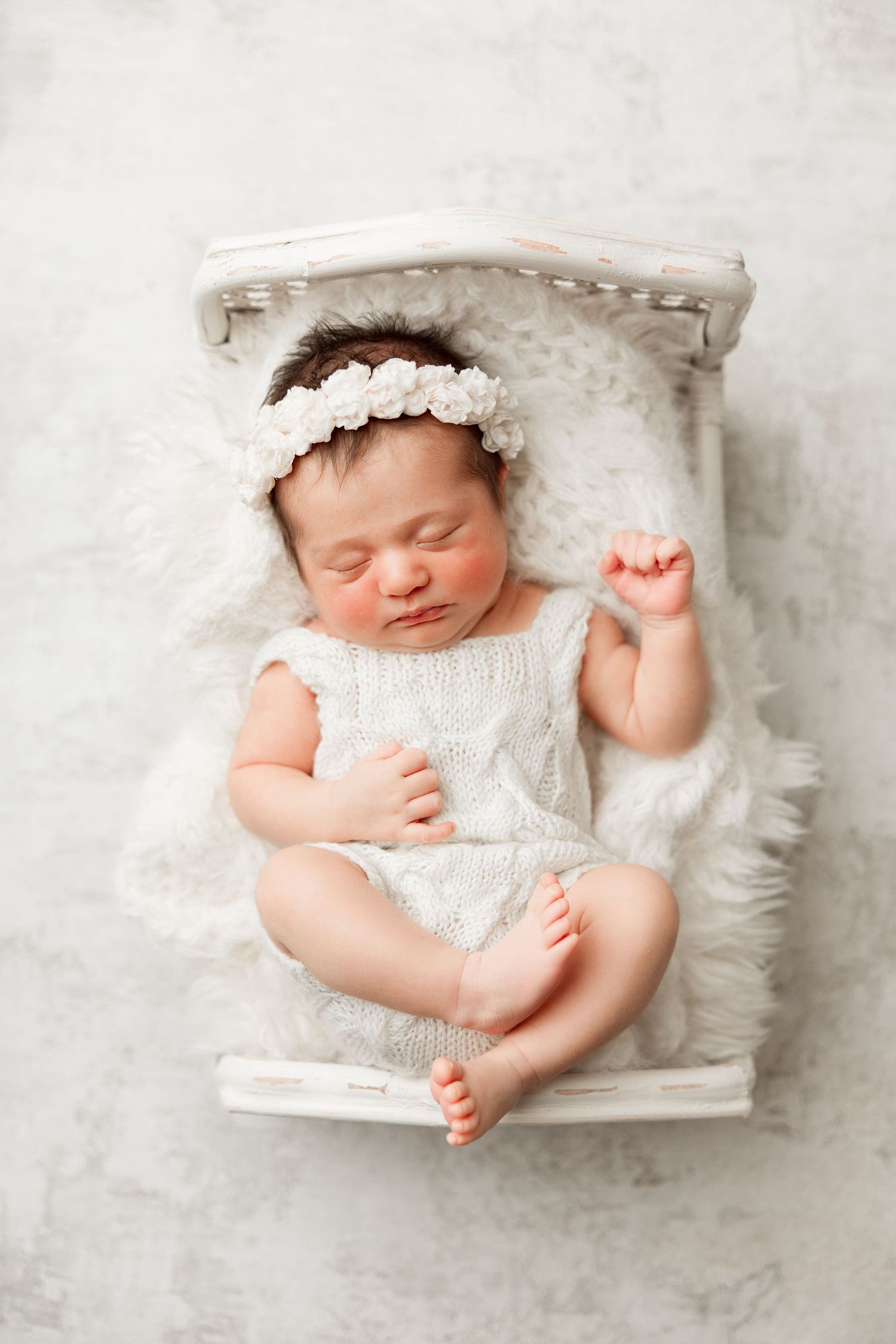 Sleeping newborn baby girl in white romper and floral headband resting on a tiny white bed with fluffy blanket.
