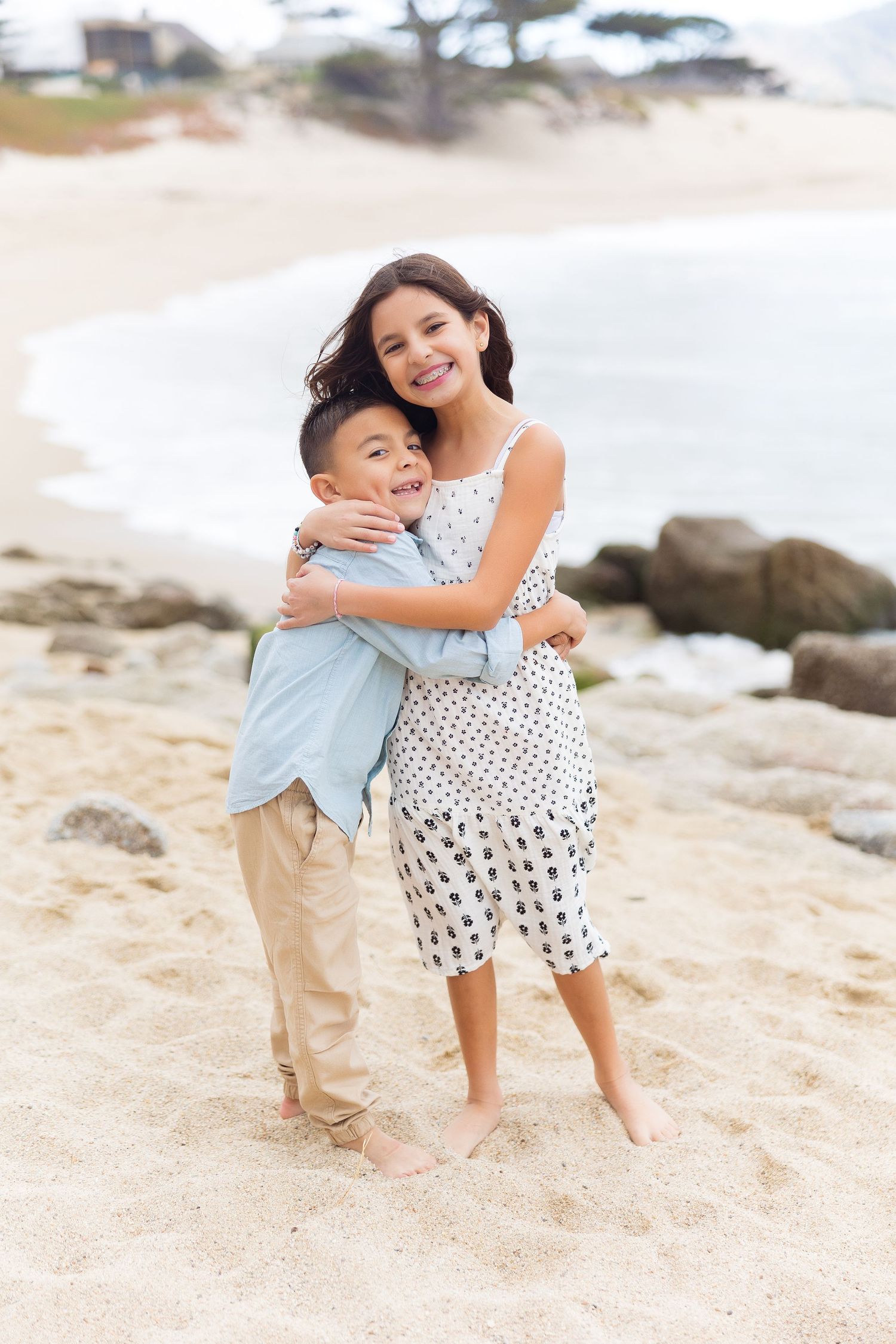 Two children embrace while standing on sandy beach with rocks and ocean.