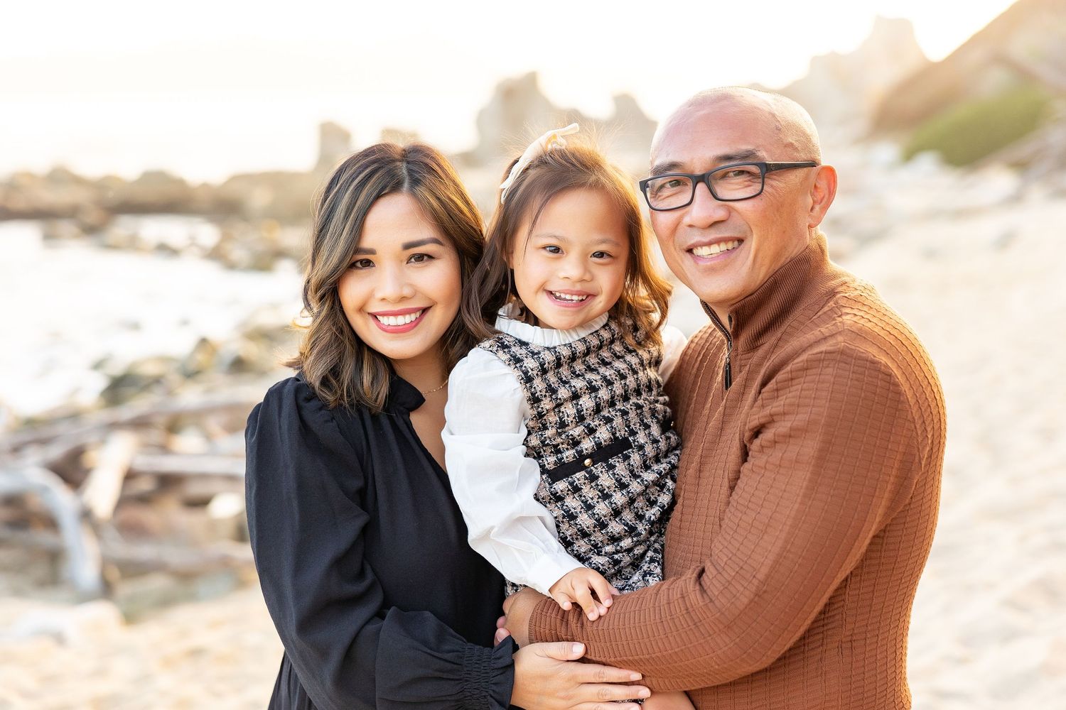Family of three posing on beach at sunset in warm toned clothing.