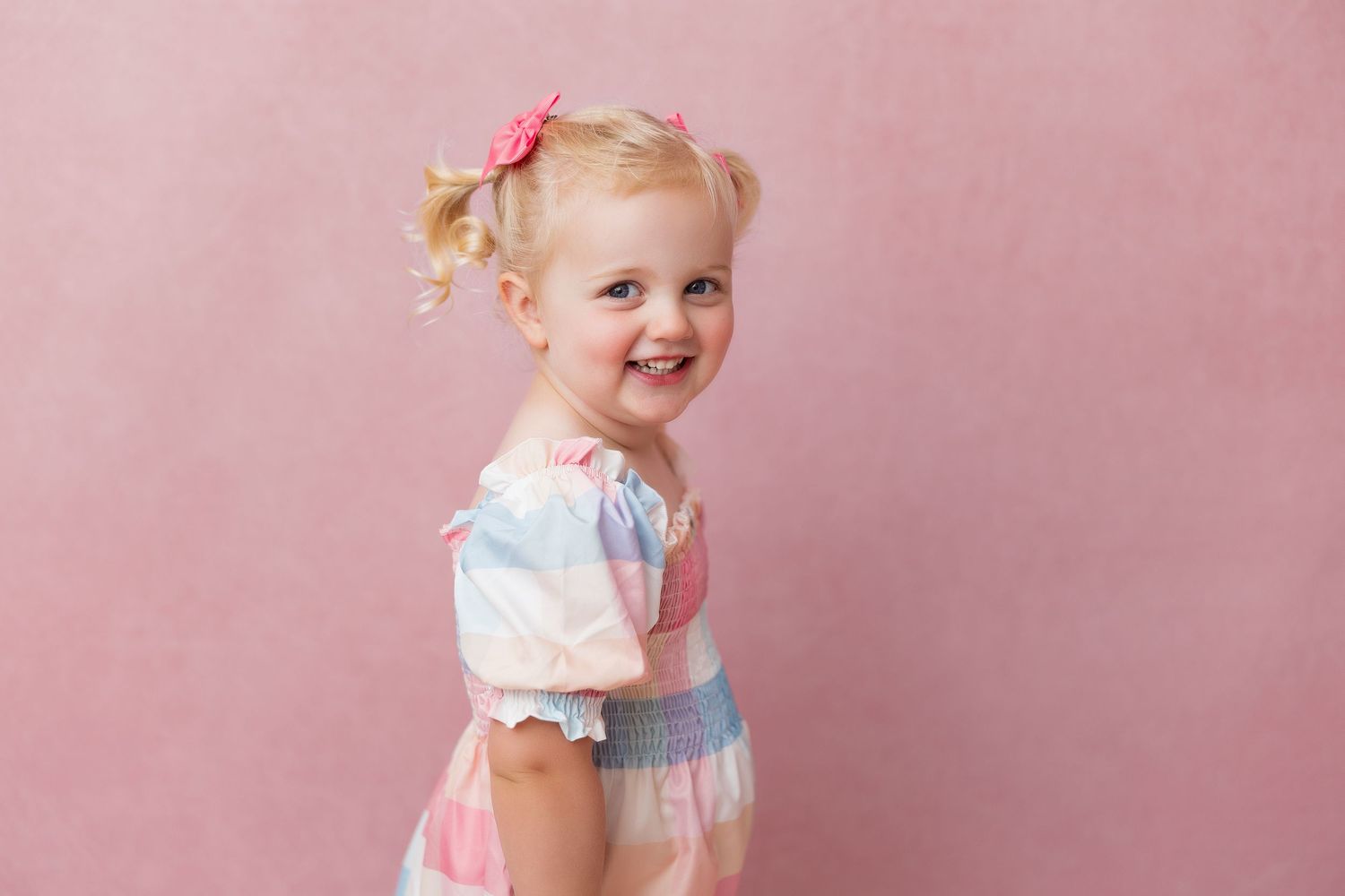 Toddler wearing a pink dress with bow detail set against a pink background for photo session.