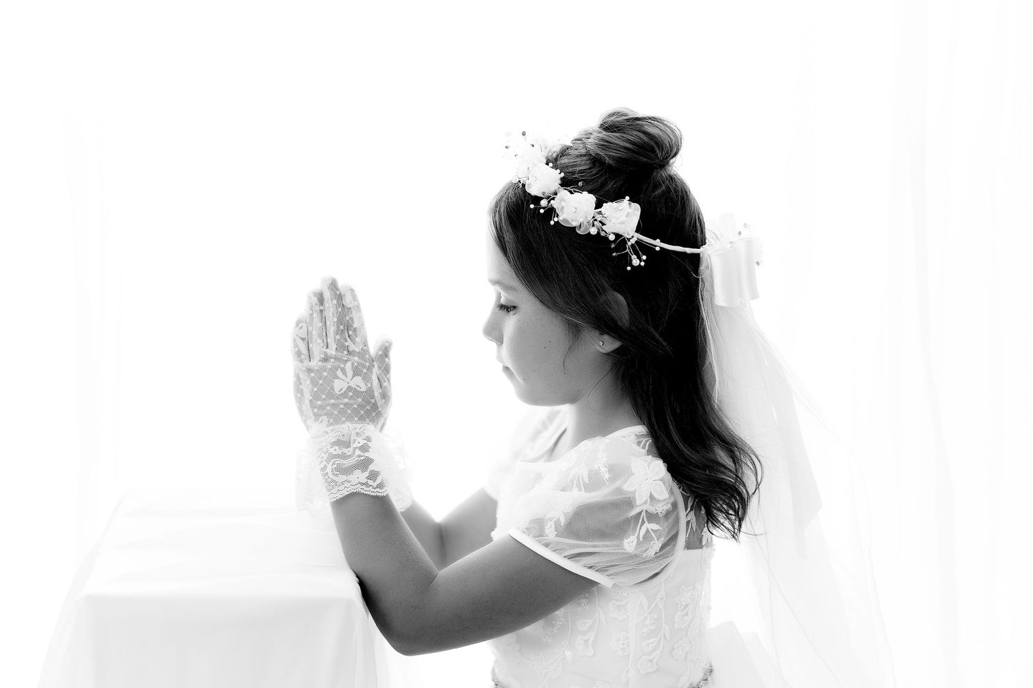 Child in white dress and veil prays with clasped hands in black and white portrait.