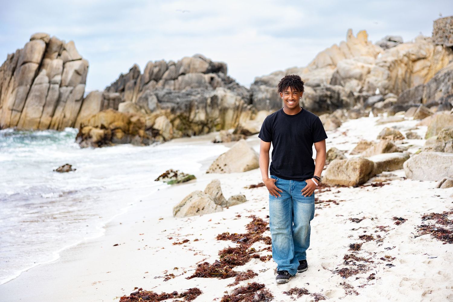 A man in a black t-shirt and jeans walks along a rocky beach with ocean waves and large boulders in the background.