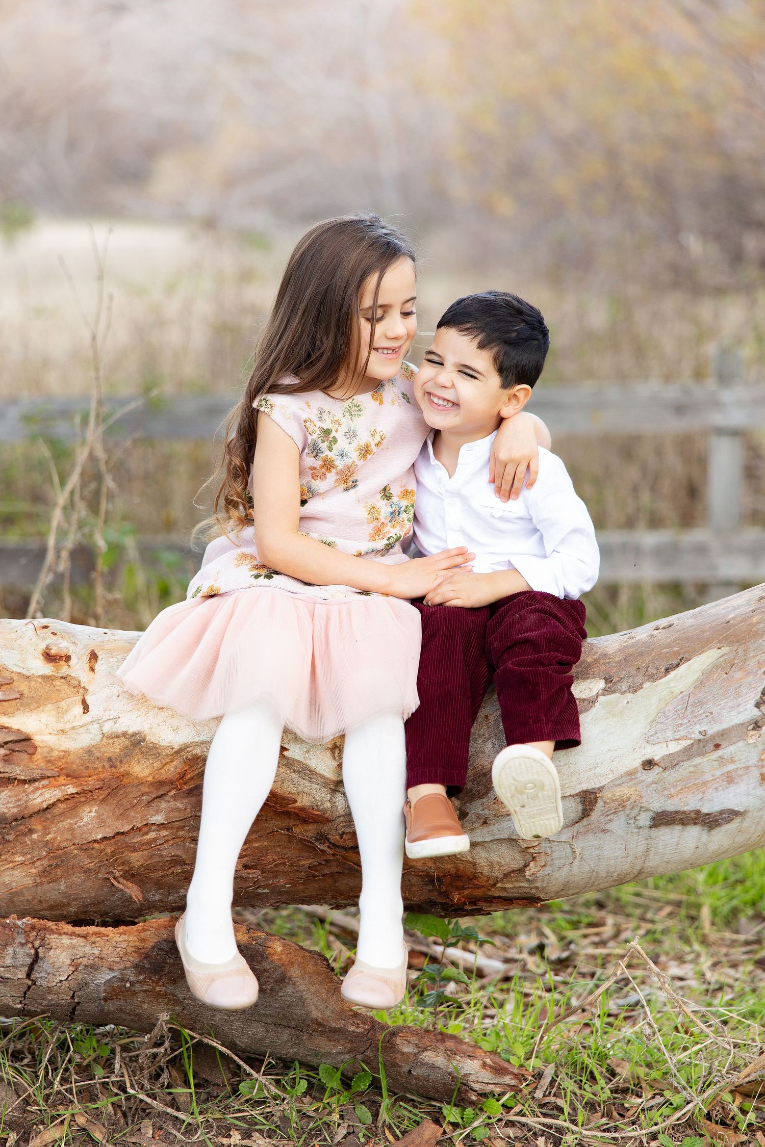 Two children sit together on a fallen log outdoors, wearing dressy clothes in a natural setting.