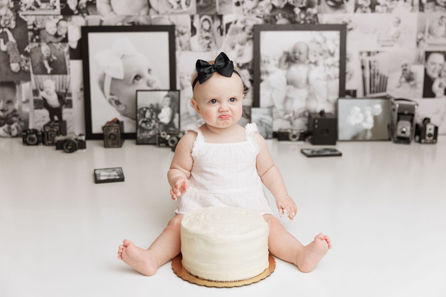 Baby wearing white outfit and black bow sitting with cake against photo collage wall.