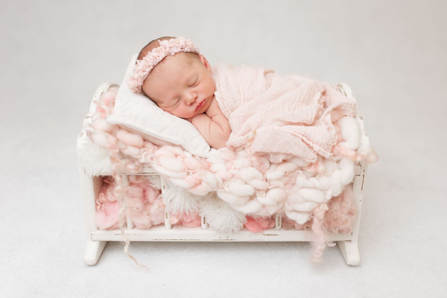 Sleeping newborn in pink outfit resting on miniature white bed with plush fabric.