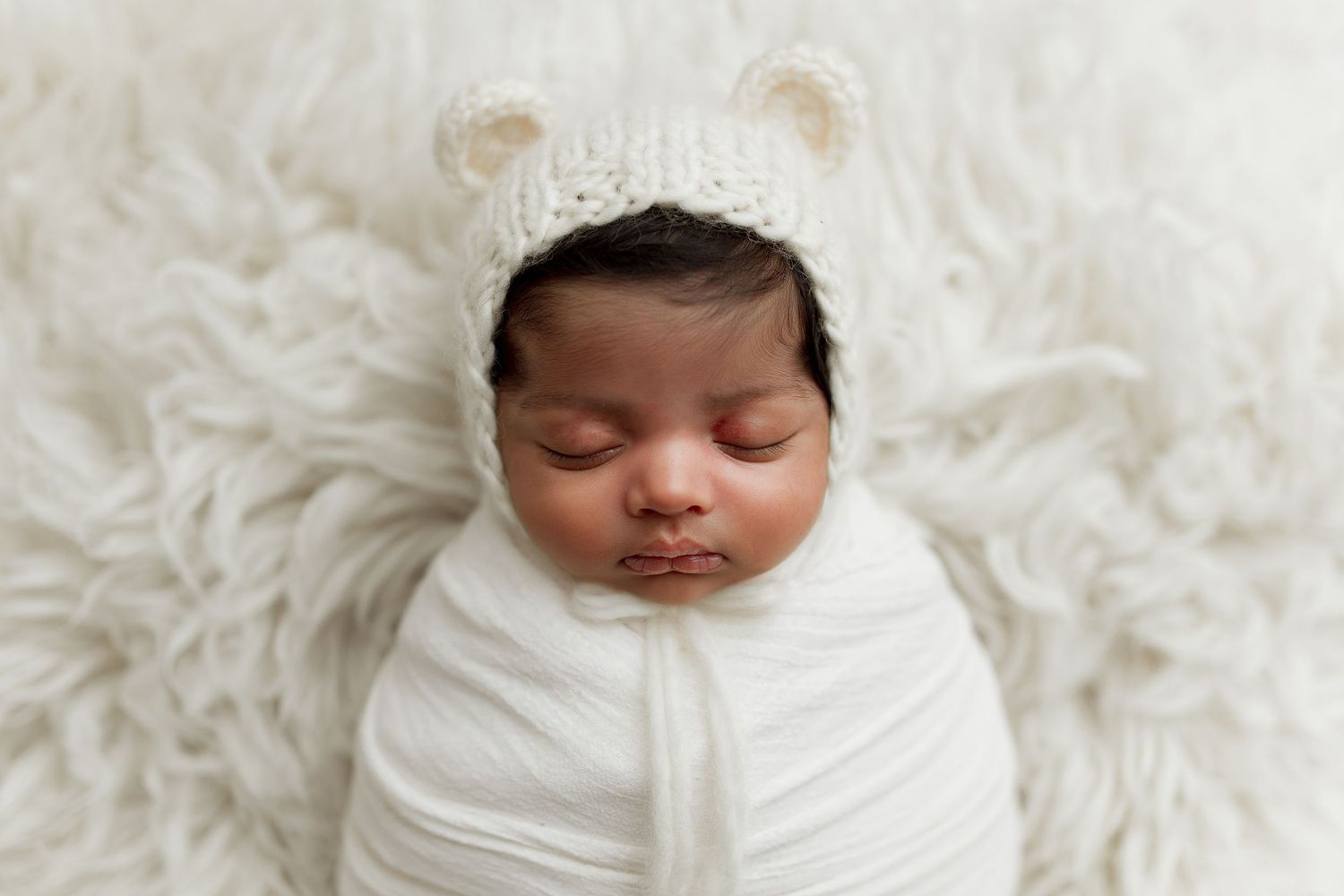 Sleeping baby wrapped in white swaddle wearing cute bear ear hat on fluffy background.